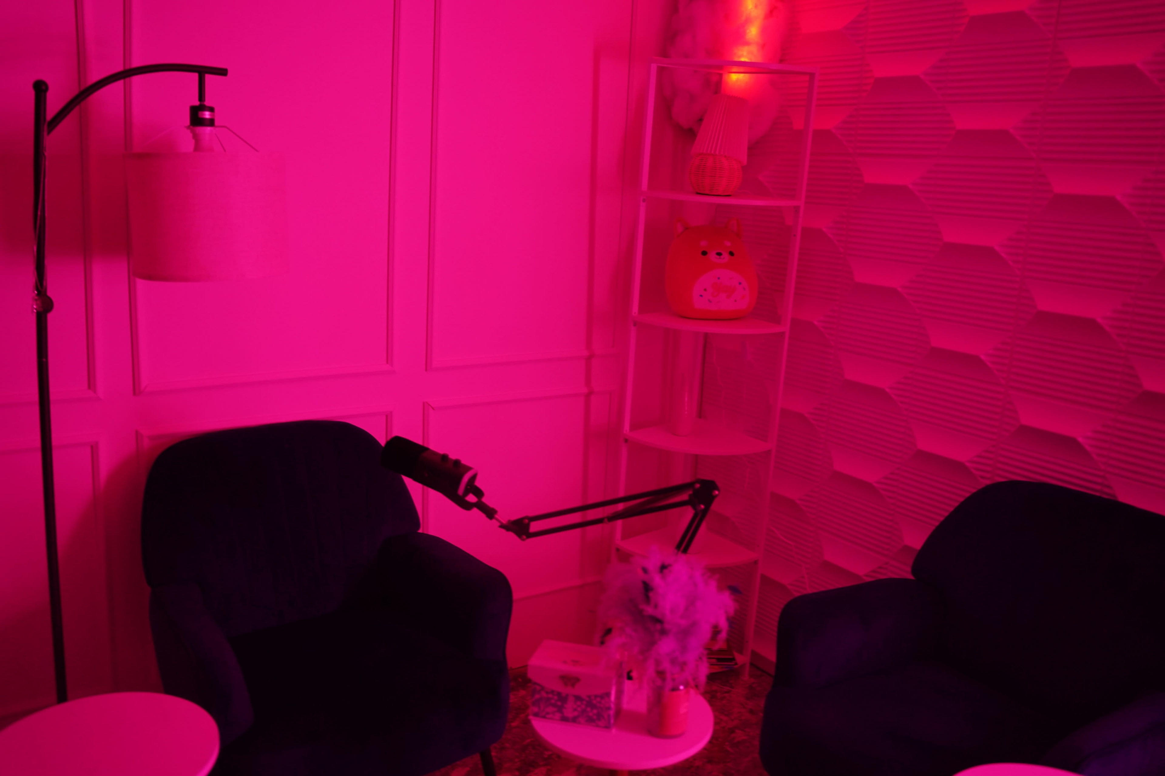 A cozy room with two dark chairs, a small white table, and a shelf displaying a decorative item, all illuminated by pink lighting.