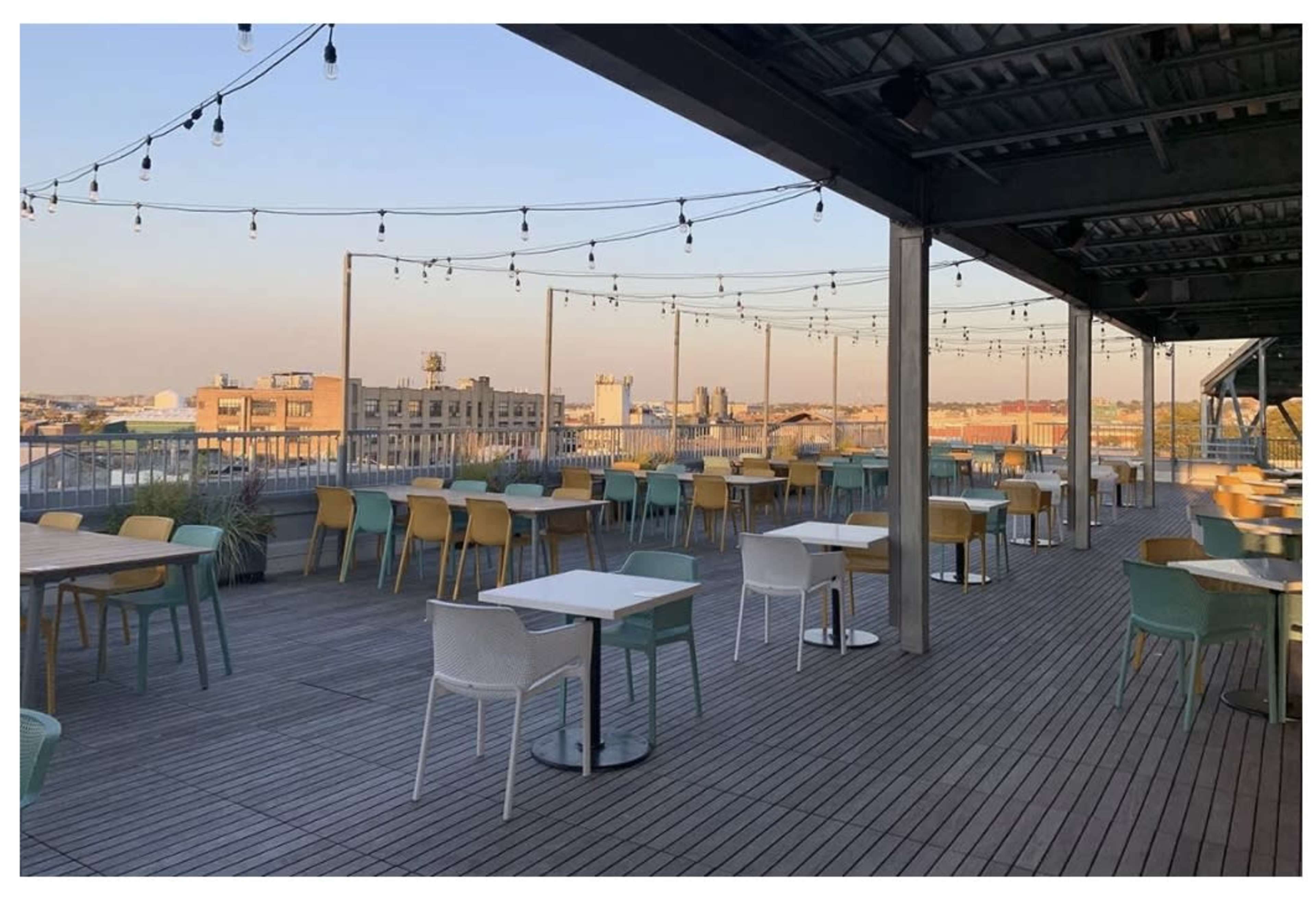 The image shows a rooftop restaurant with empty tables and chairs arranged on a wooden deck, illuminated by string lights against a clear evening sky.