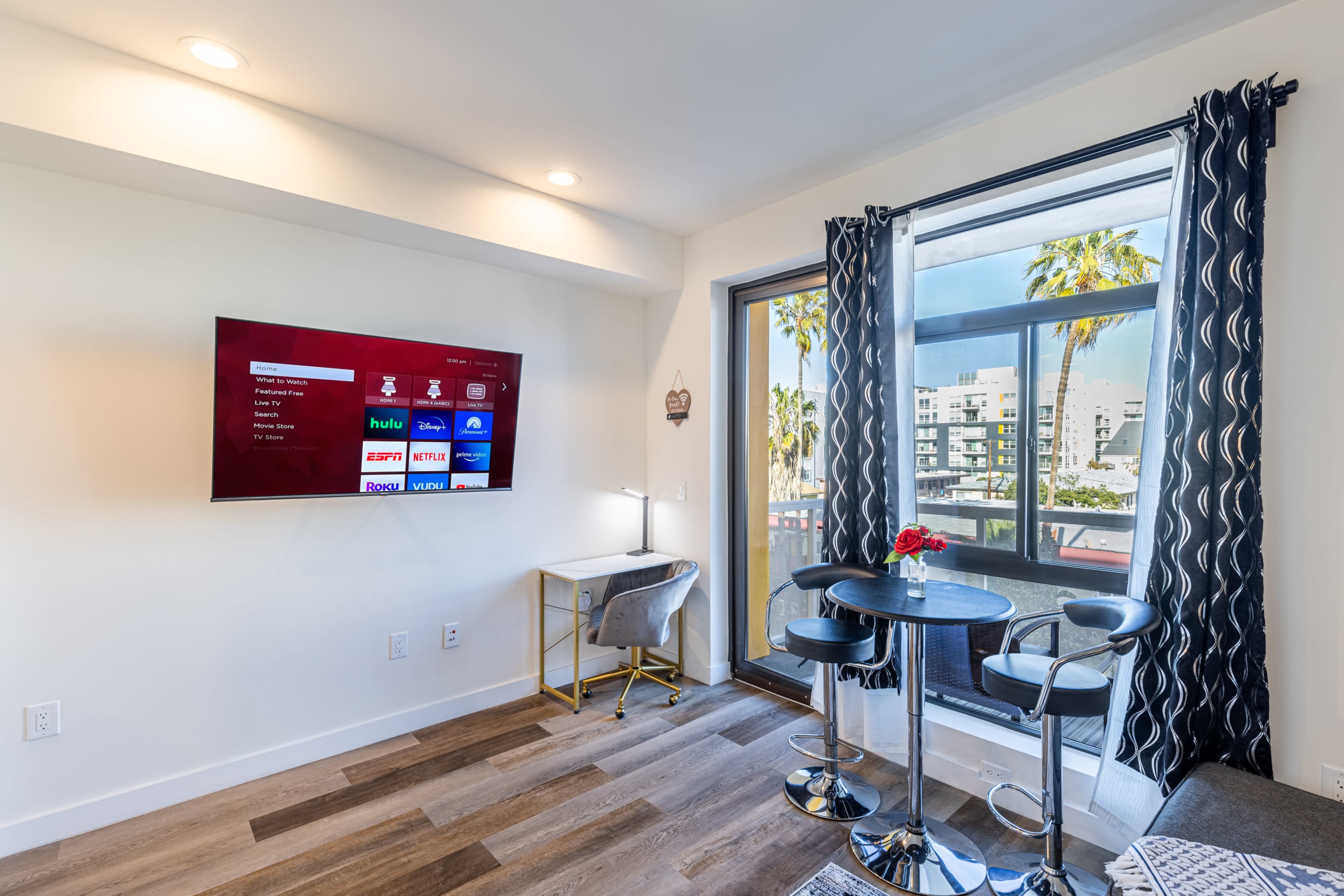 A modern living space features a flat-screen TV mounted on the wall, a small dining table with two chairs, and large windows overlooking palm trees.