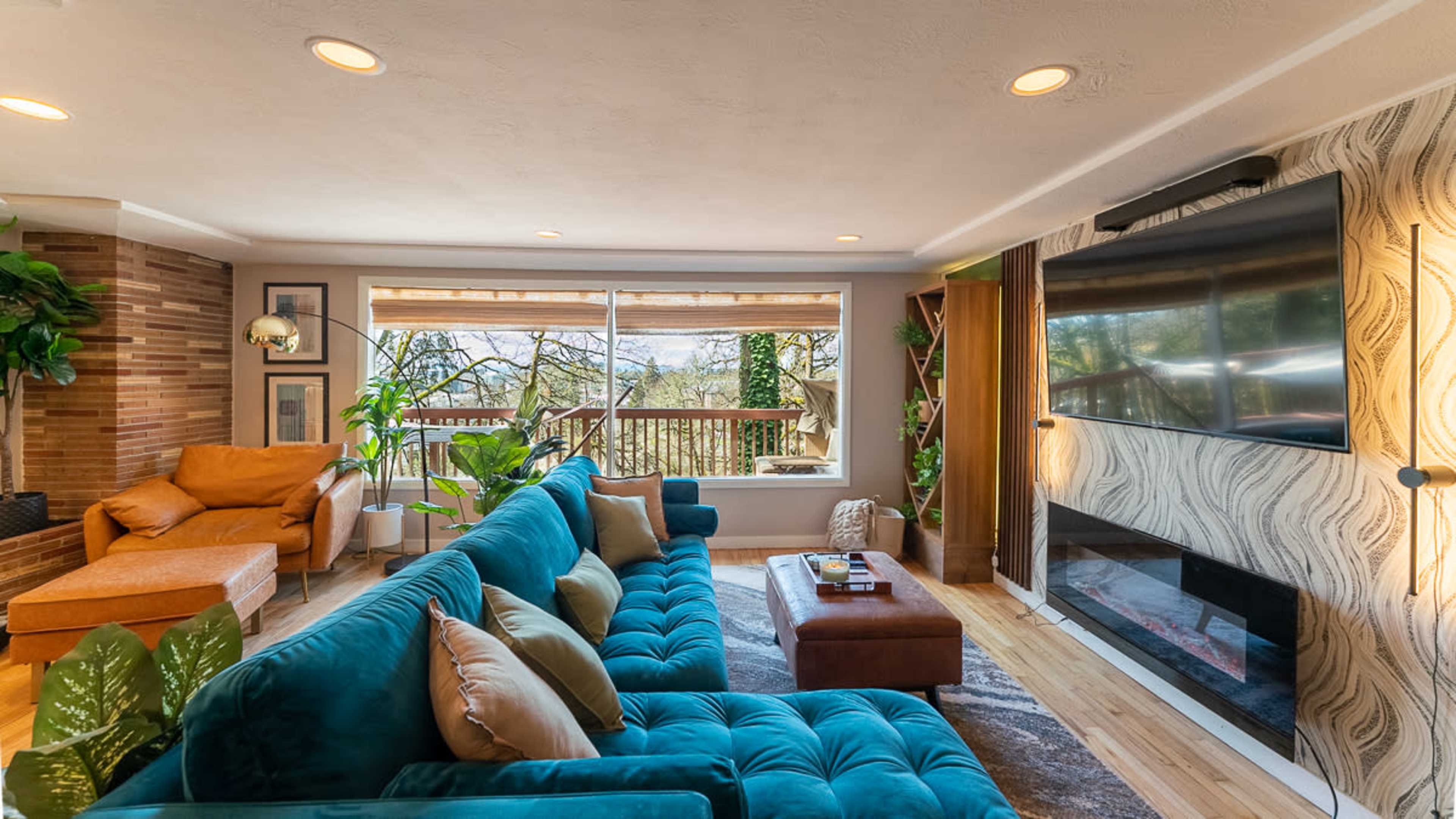 A modern living room features a large turquoise sofa, a brown armchair, and a glass door leading to a deck with views of trees.