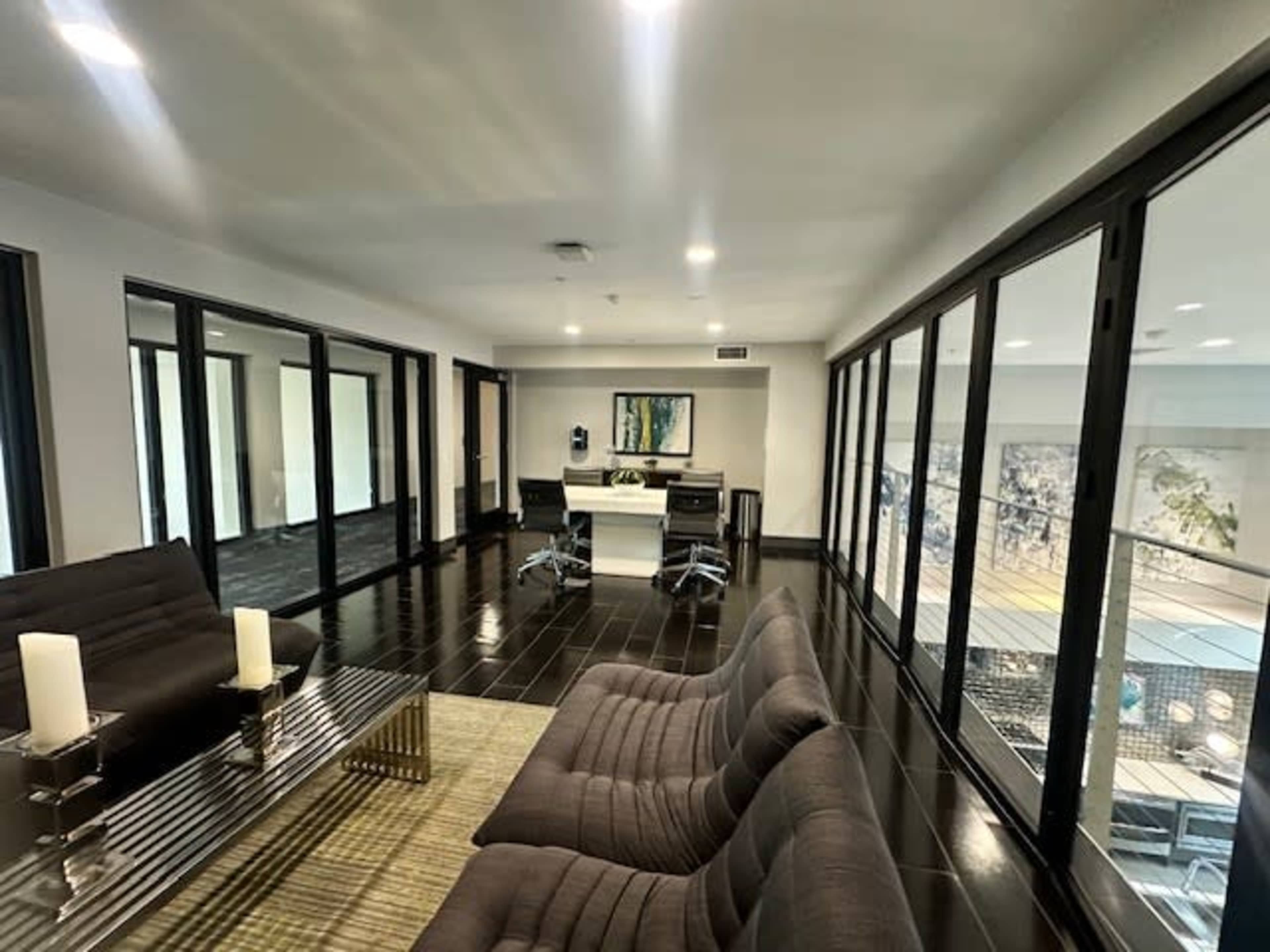 The image shows a modern office space with large glass panels, a sleek table, and two chairs arranged around a coffee table.