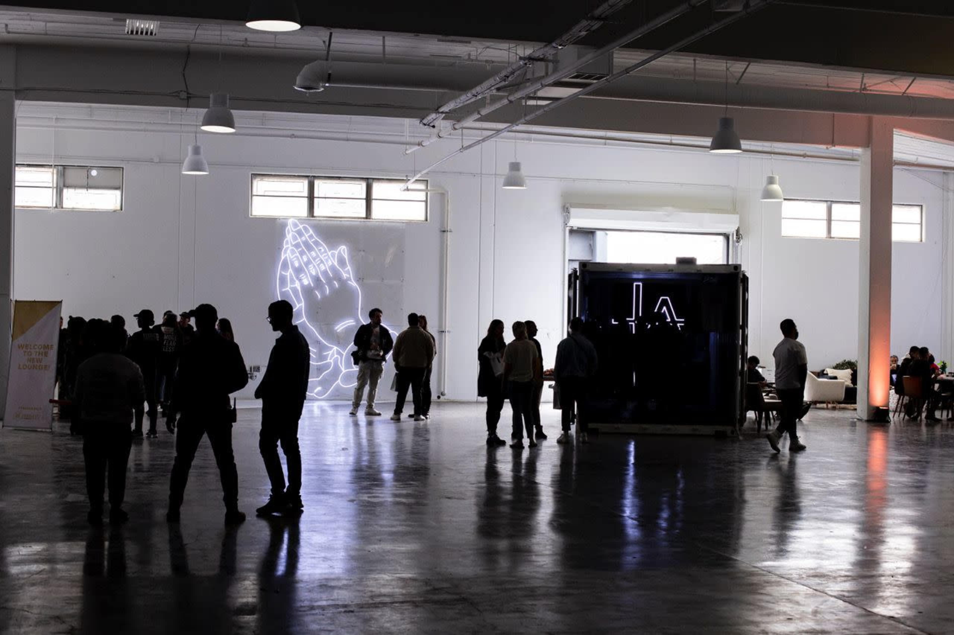 A spacious indoor venue with a large neon artwork of clasped hands on the wall and several groups of people chatting in the foreground.