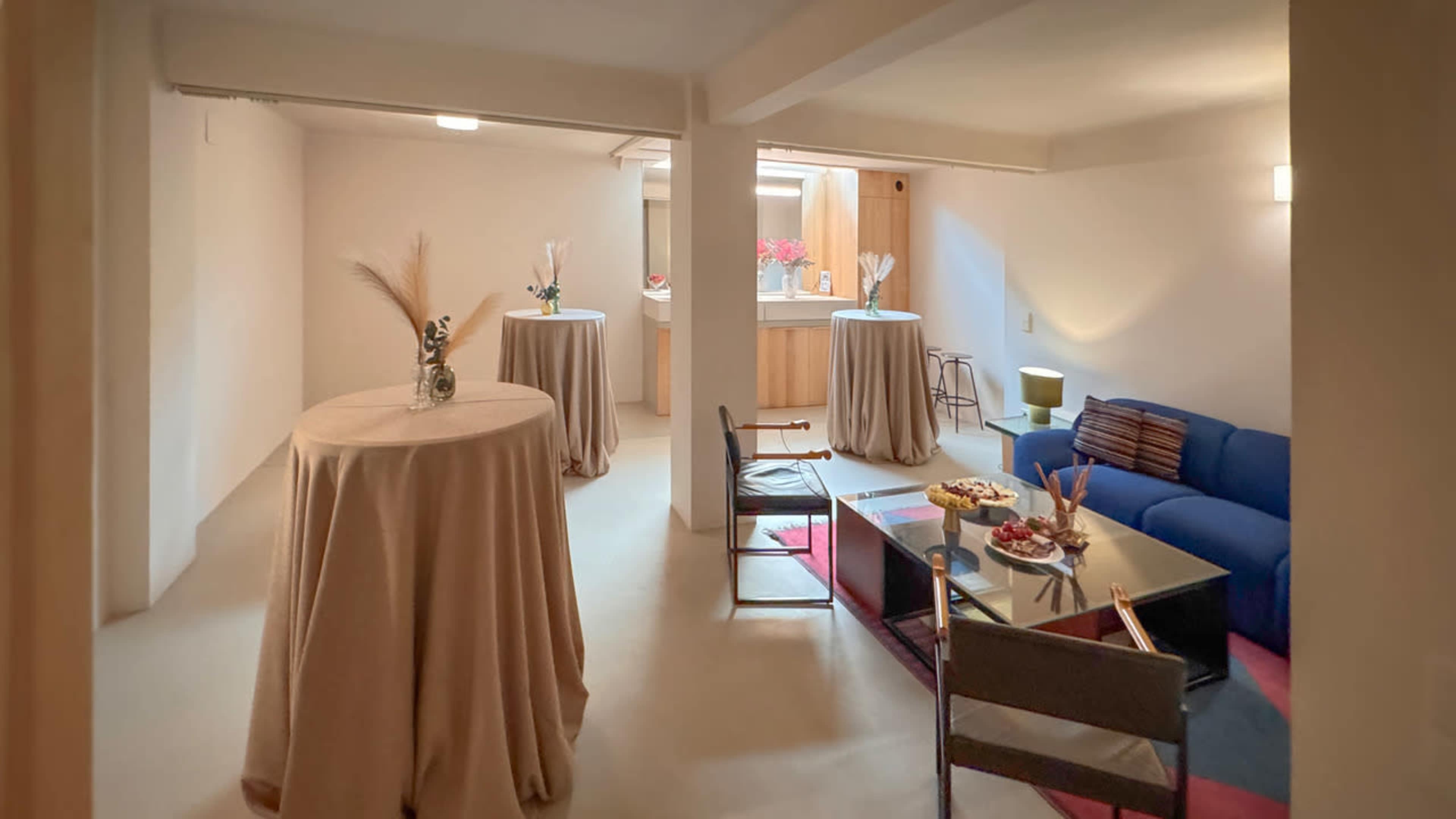 The image shows a minimalistic interior space with a blue sofa, a glass coffee table, and tables covered in beige cloth, leading to a kitchen area in the background.