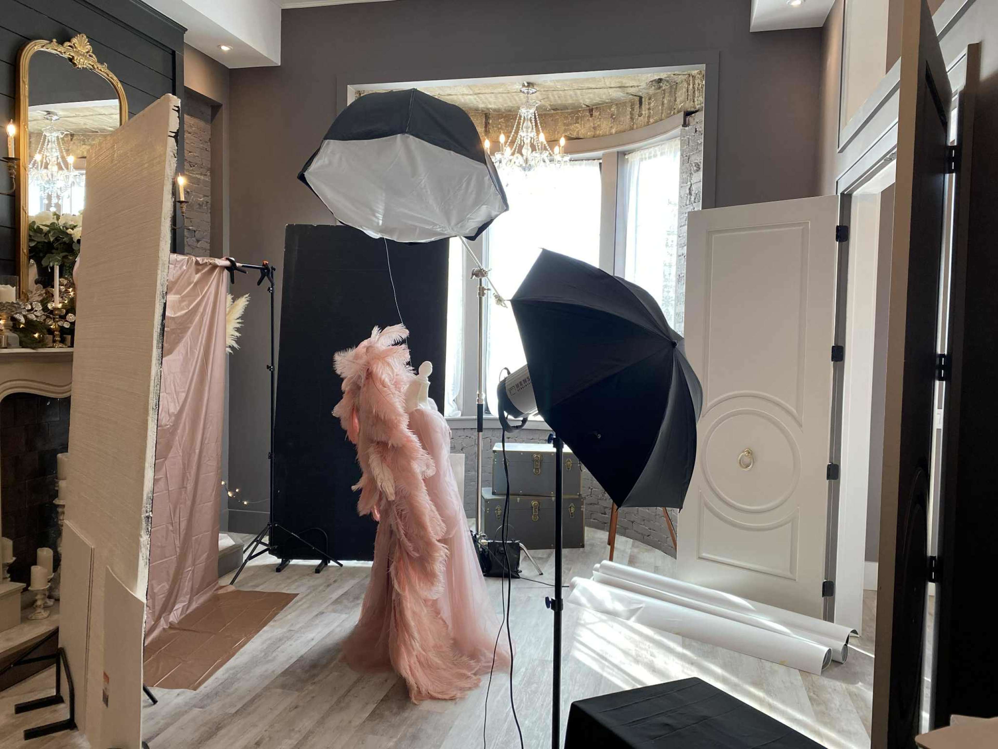 A model in a pink gown poses in a well-lit photo studio with backdrops and lighting equipment.