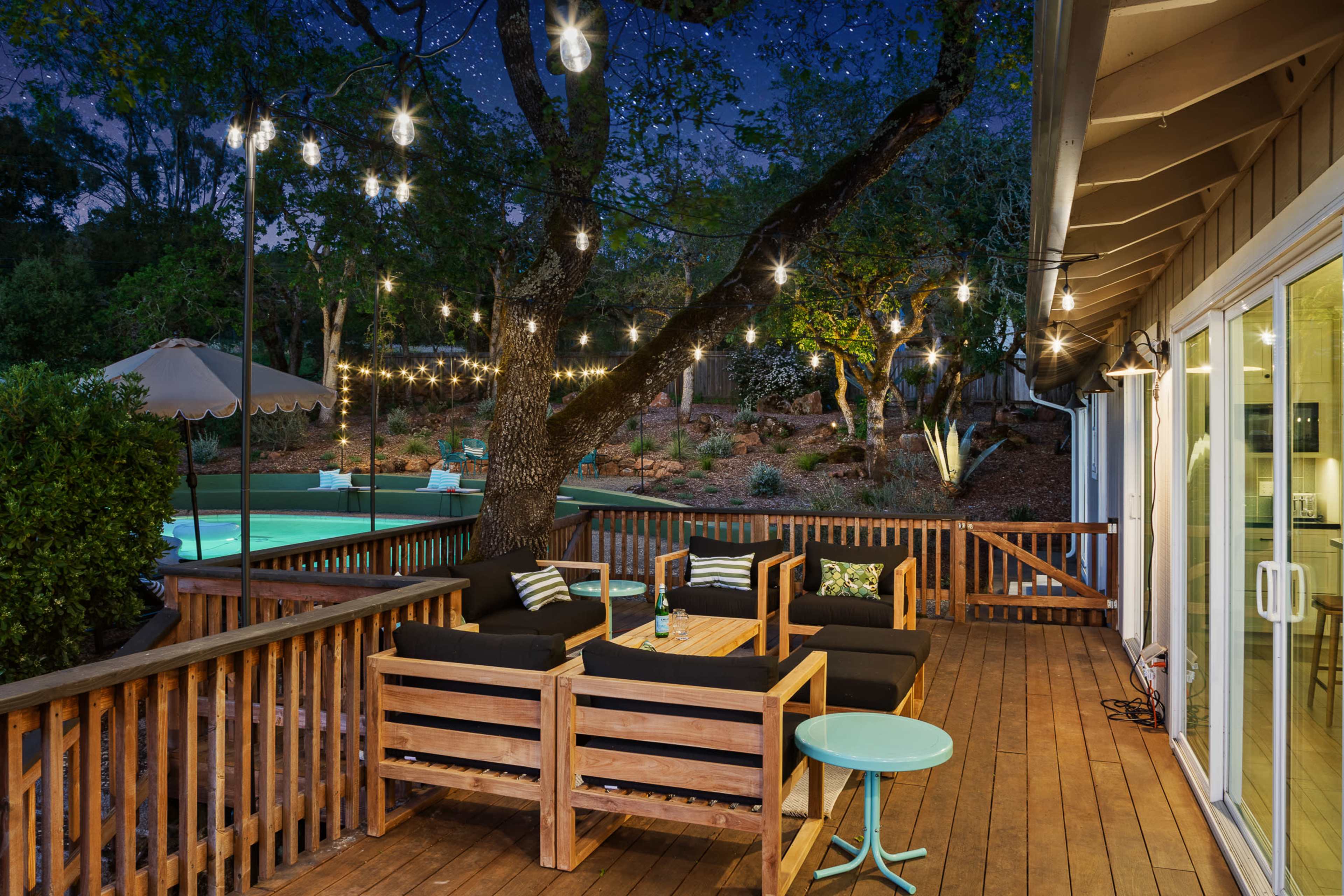 A deck area with wooden furniture, string lights overhead, and a view of a pool surrounded by greenery under a night sky.