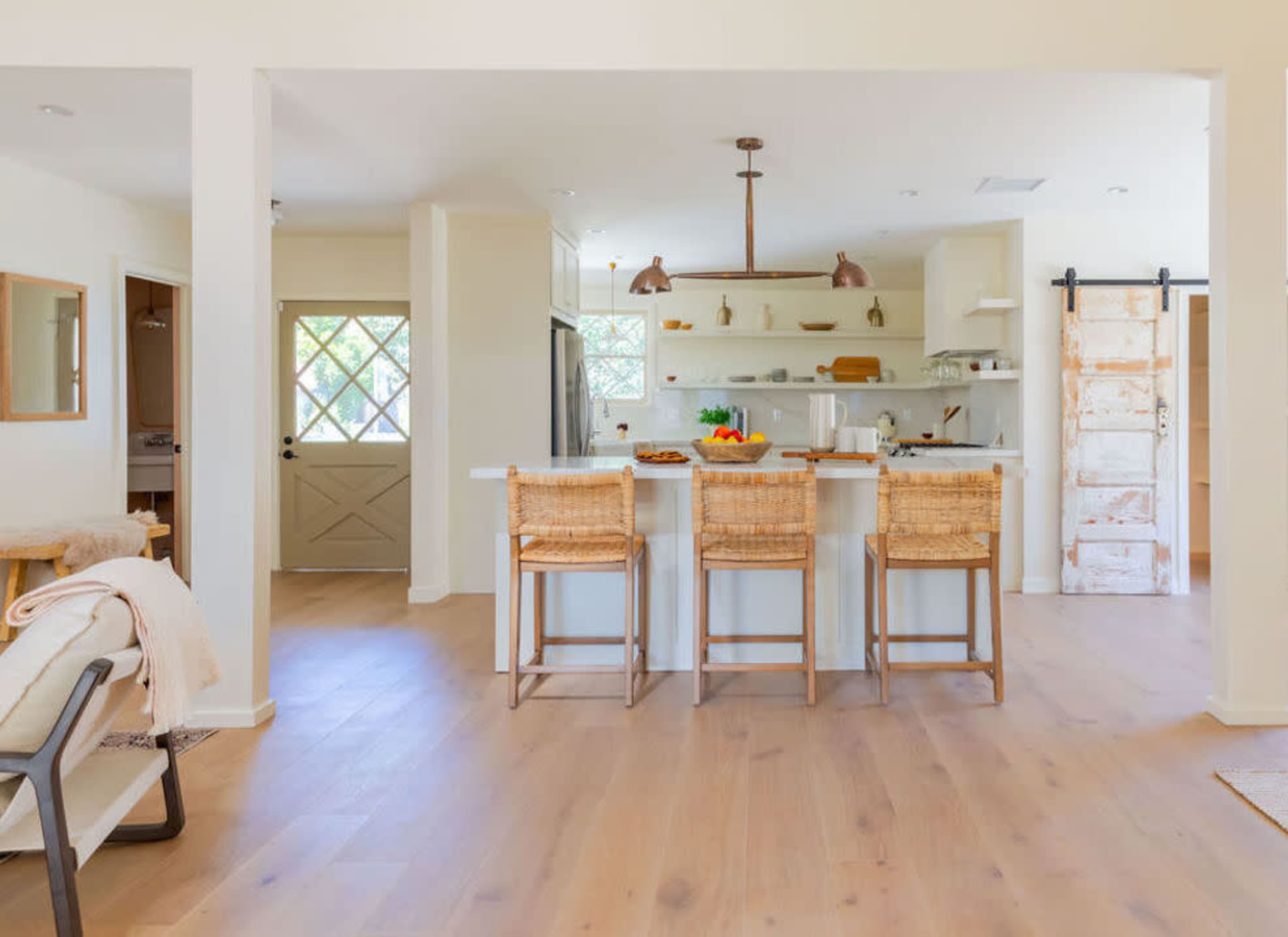 A bright, modern kitchen features a central island with three woven bar stools, open shelving, and a door leading to an entrance area.