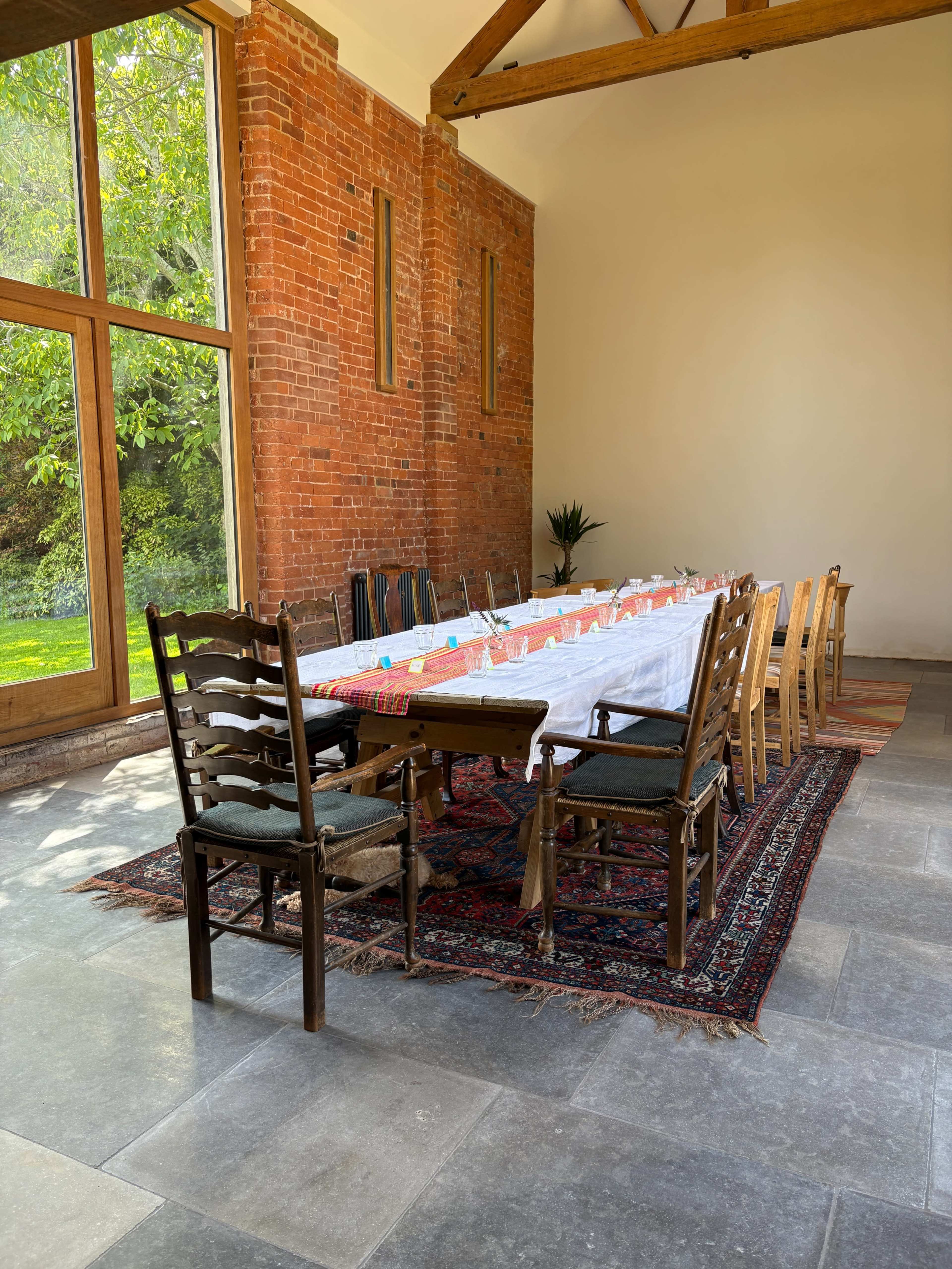 A long dining table is set with white and colorful tableware in a well-lit room with large windows and exposed brick walls.
