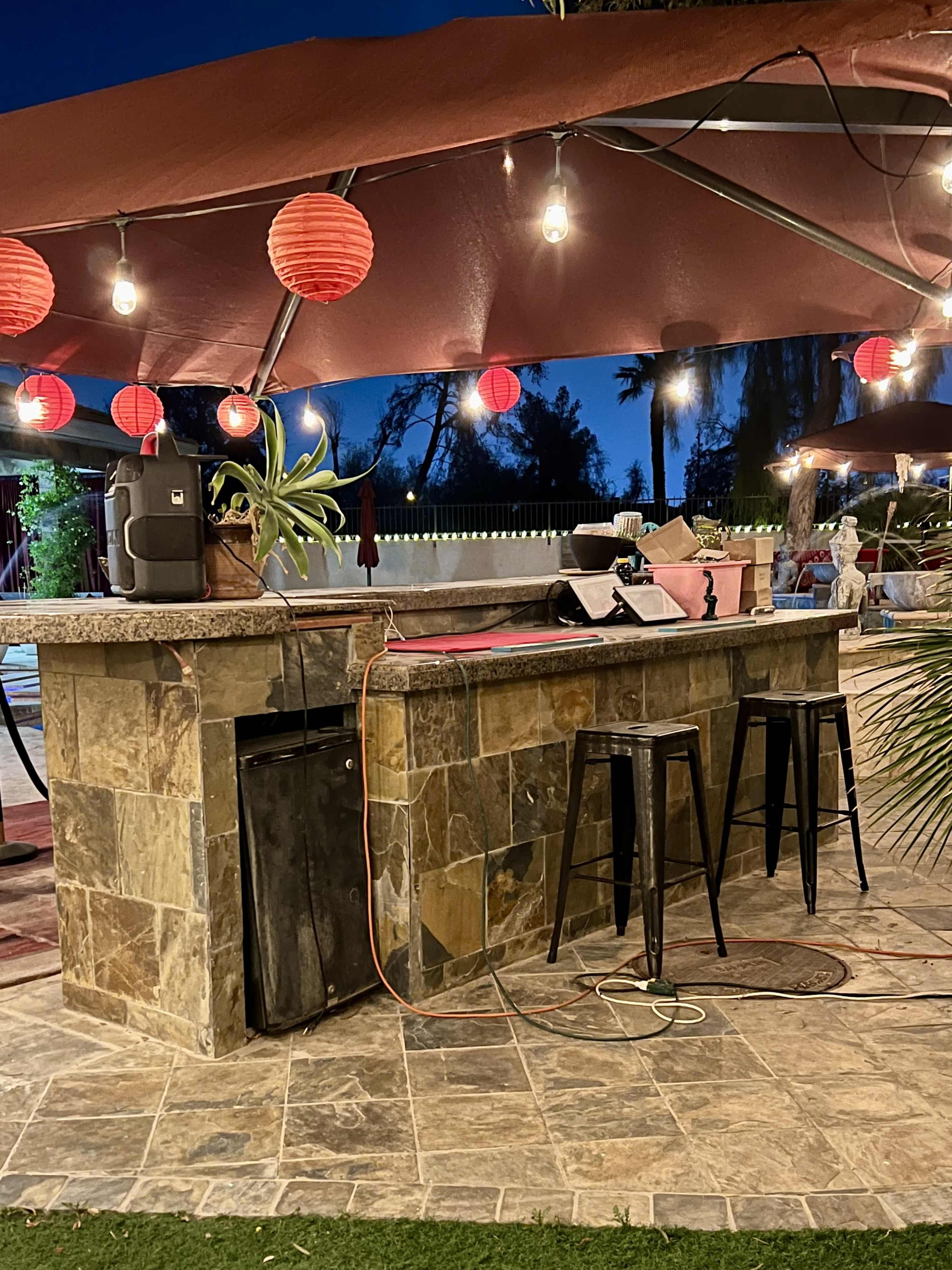 The scene features an outdoor bar area illuminated by red lanterns and string lights, with a stone countertop and high stools, set against a twilight backdrop.