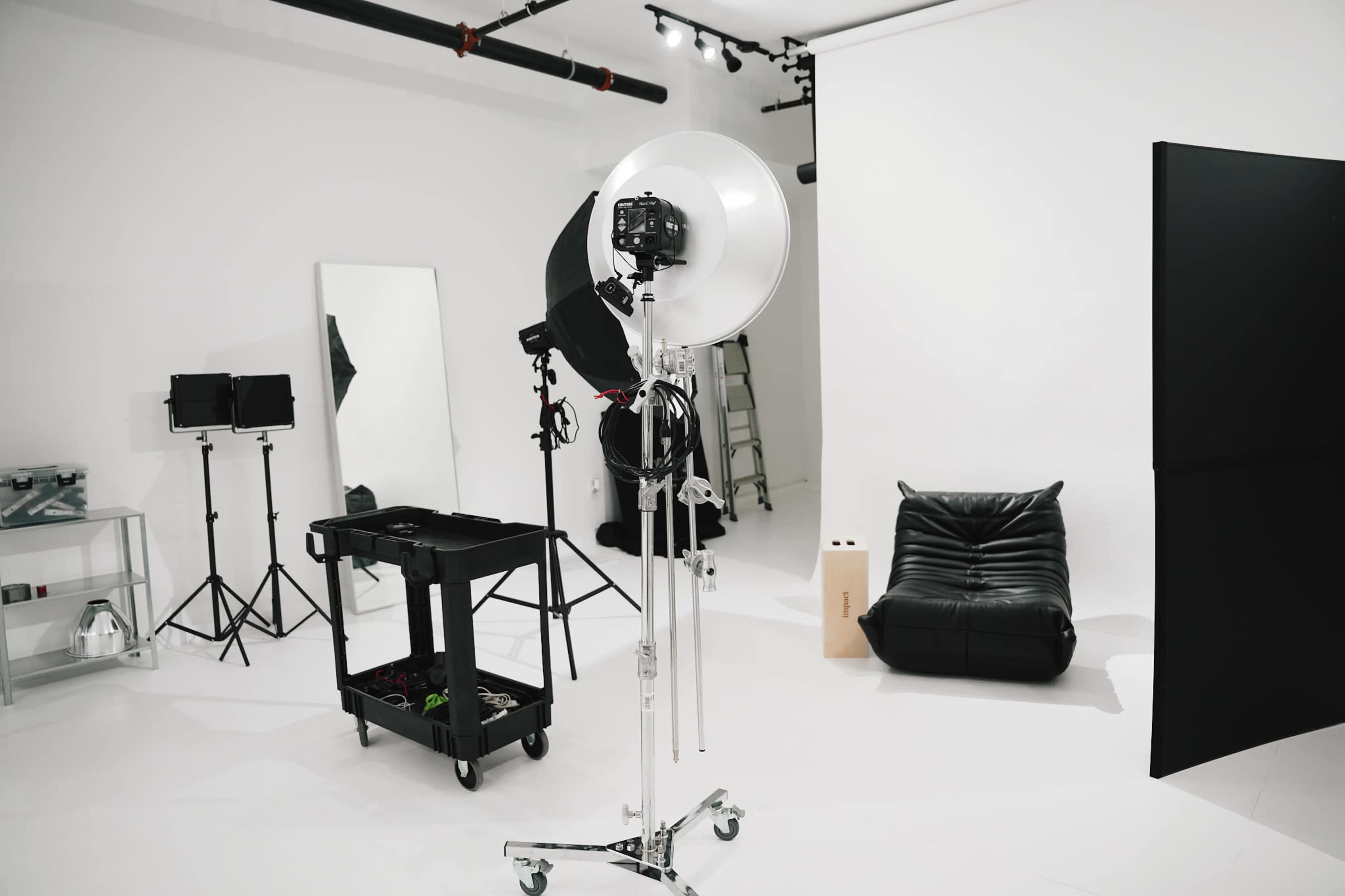A photography studio is set up with a circular light, a camera on a stand, a black couch, a large mirror, and various equipment arranged on carts.