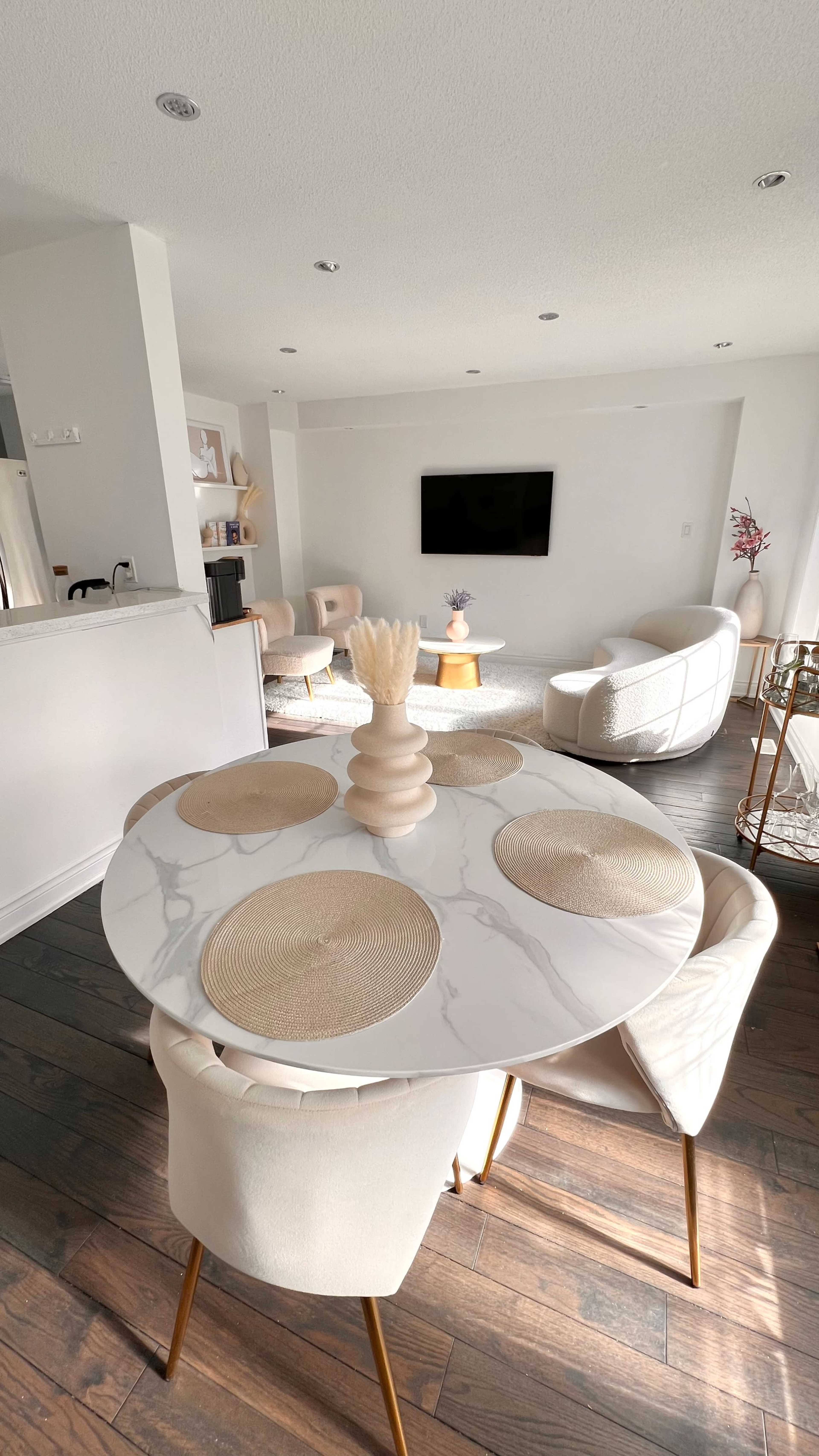 A modern dining area features a round marble table with beige placemats and four upholstered chairs, leading into a bright living room with a television and soft seating.