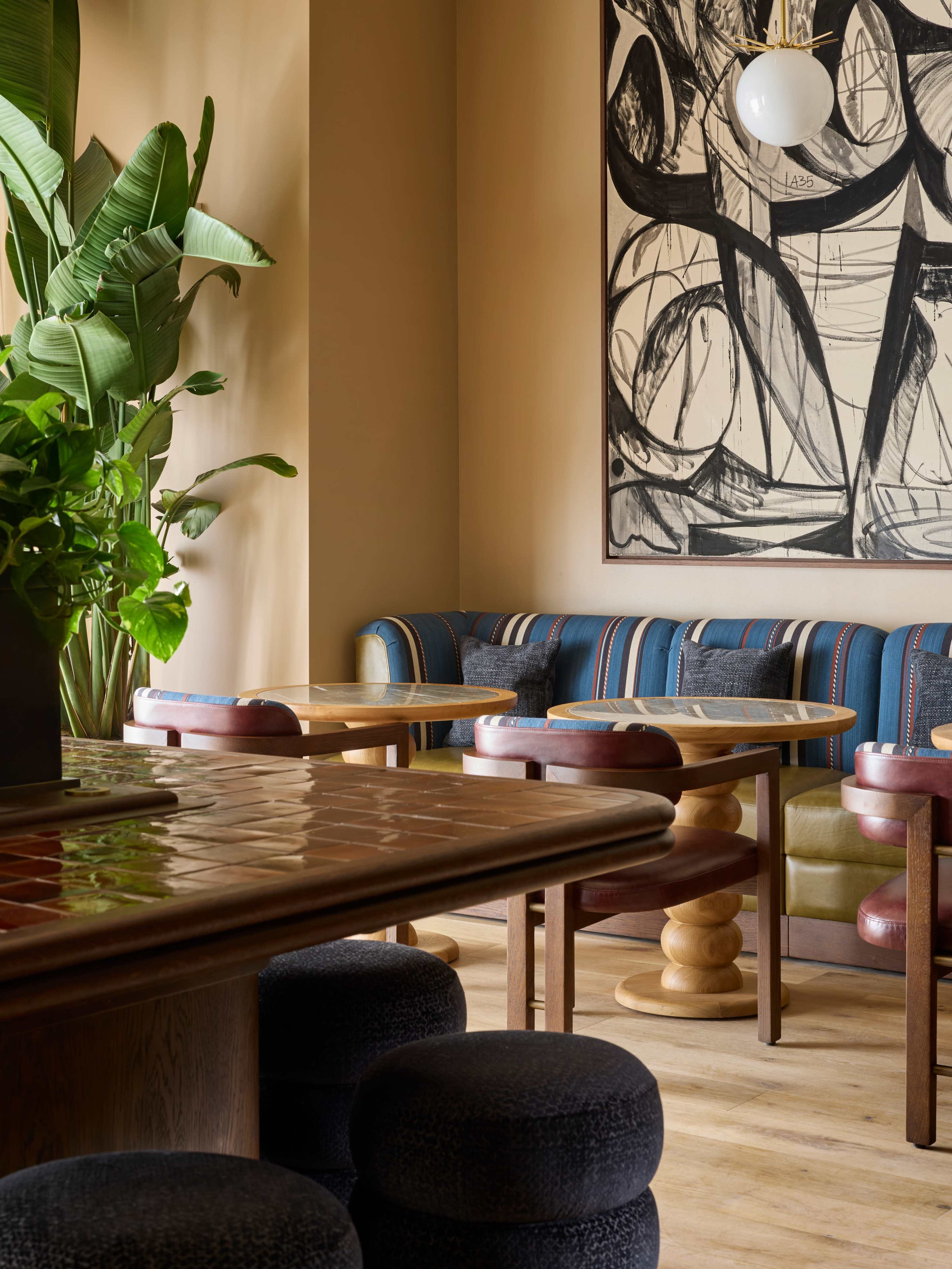 The image depicts a stylish café interior featuring wooden tables, upholstered seating, a large abstract painting on the wall, and a lush plant in the corner.