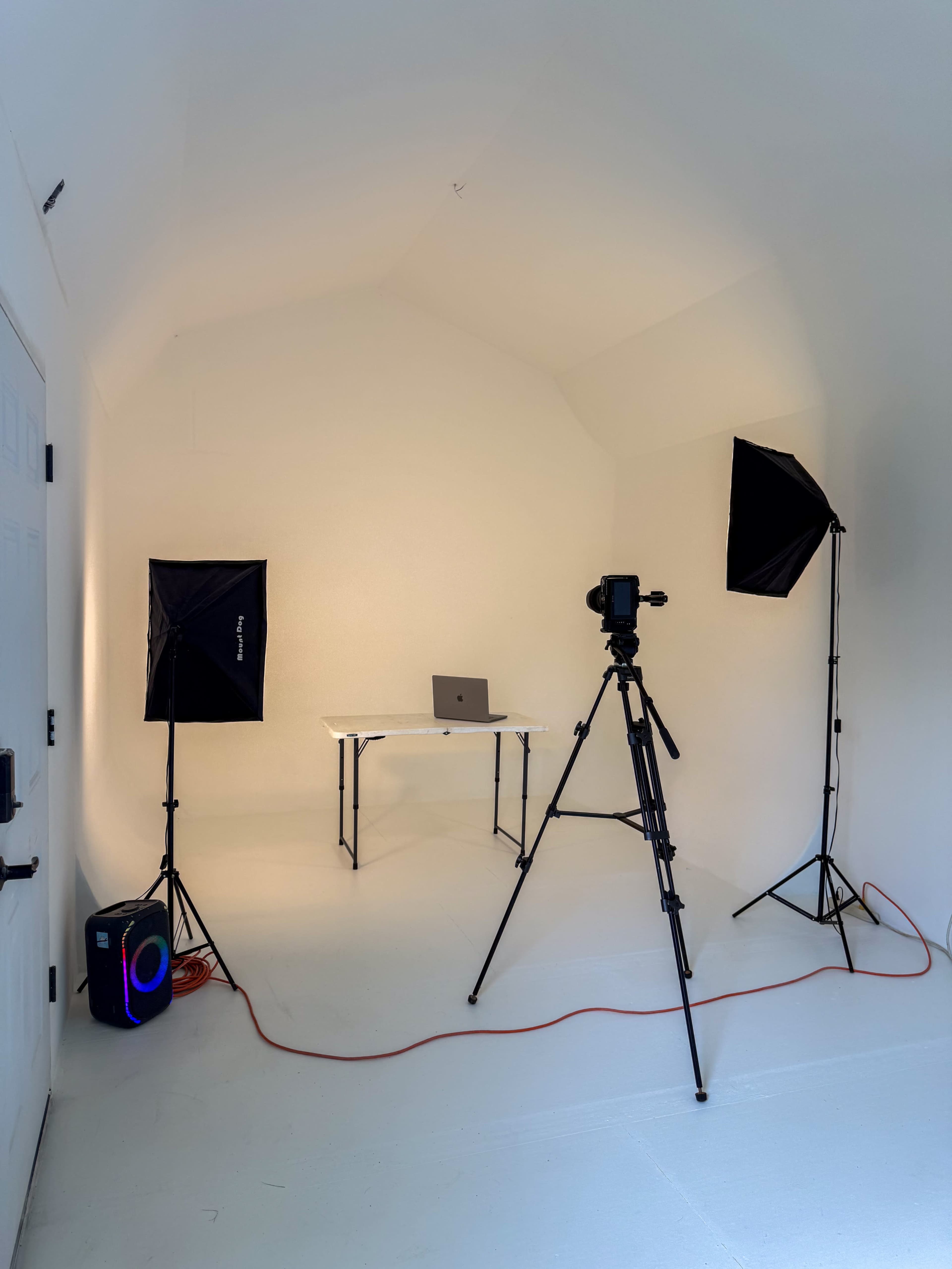 The image shows a minimalist photo studio with a table and a laptop, flanked by two softbox lights and a tripod.