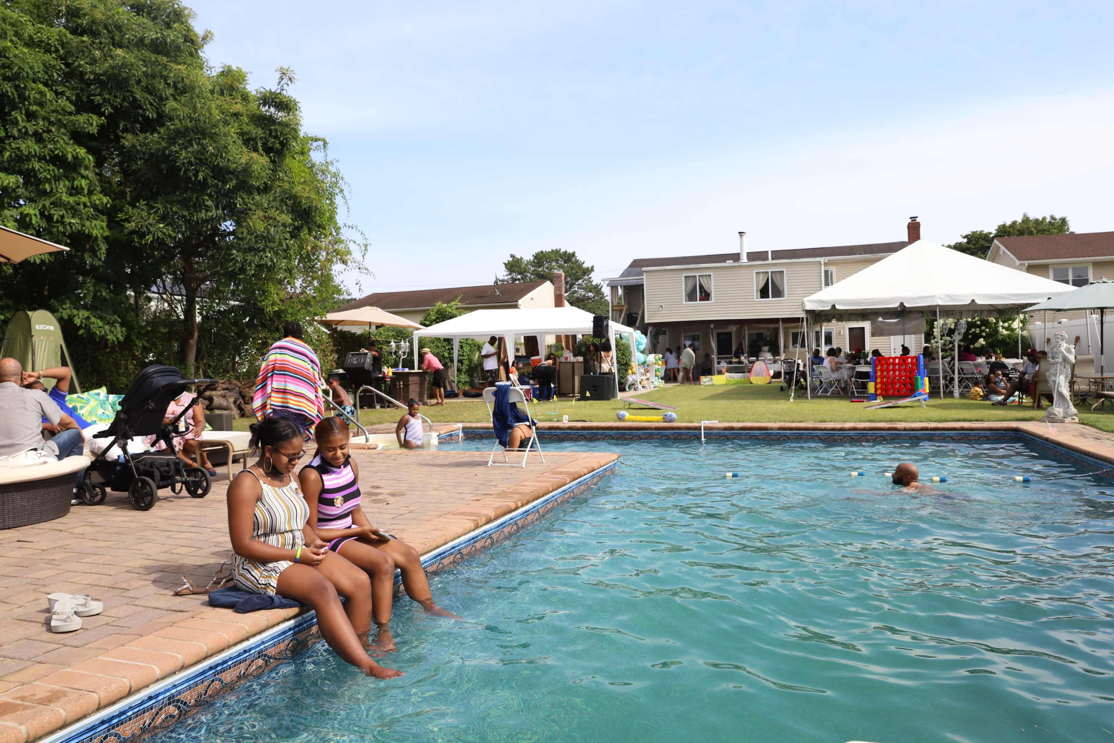 A woman and a girl sit on the edge of a swimming pool while several people socialize nearby under tents and umbrellas in a backyard setting.