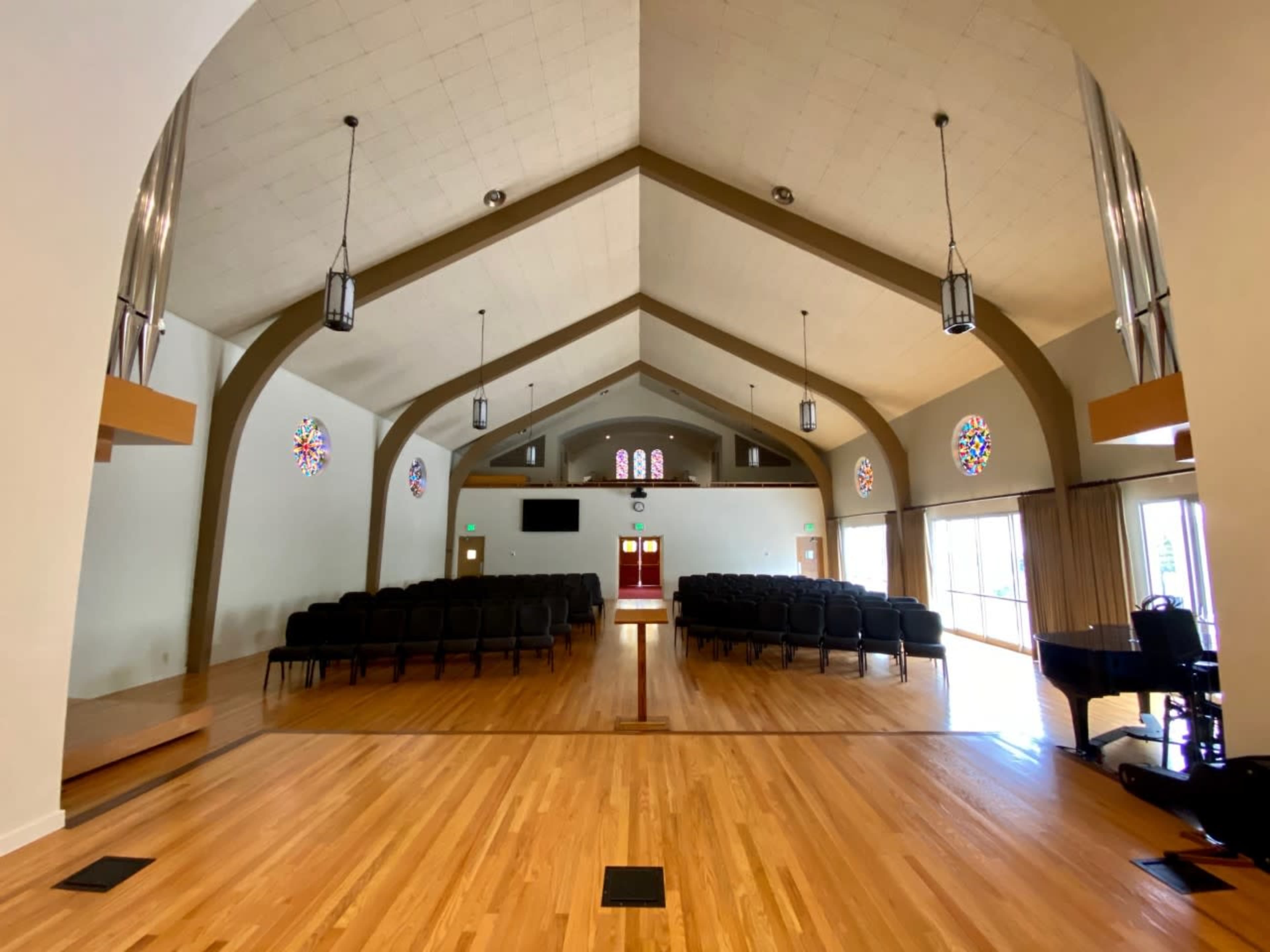 A spacious, well-lit interior of a church with rows of black chairs, stained glass windows, and a polished wooden floor.