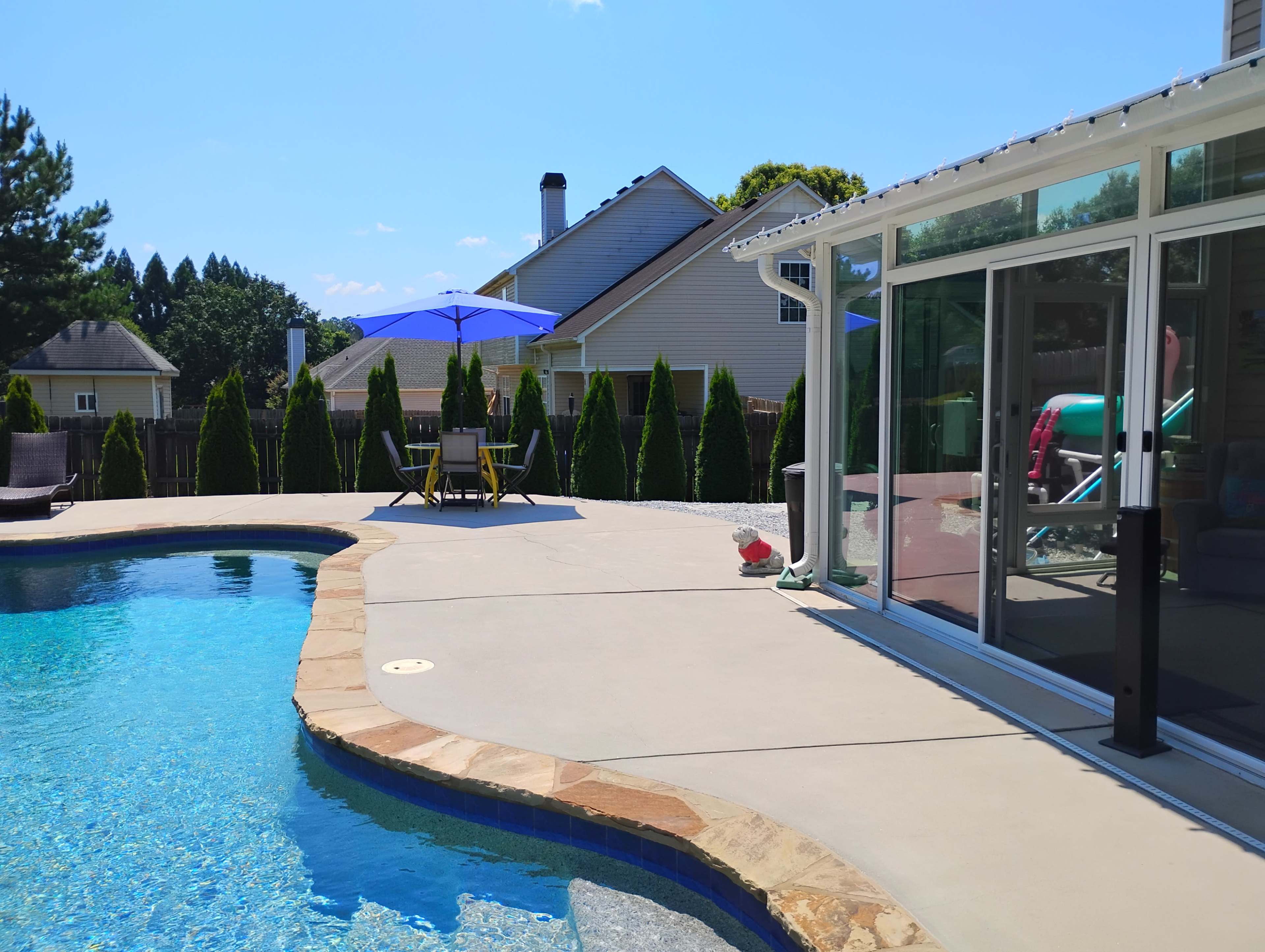 The image shows a swimming pool beside a patio with a lounge chair and a blue umbrella, surrounded by tall green hedges and a house in the background.
