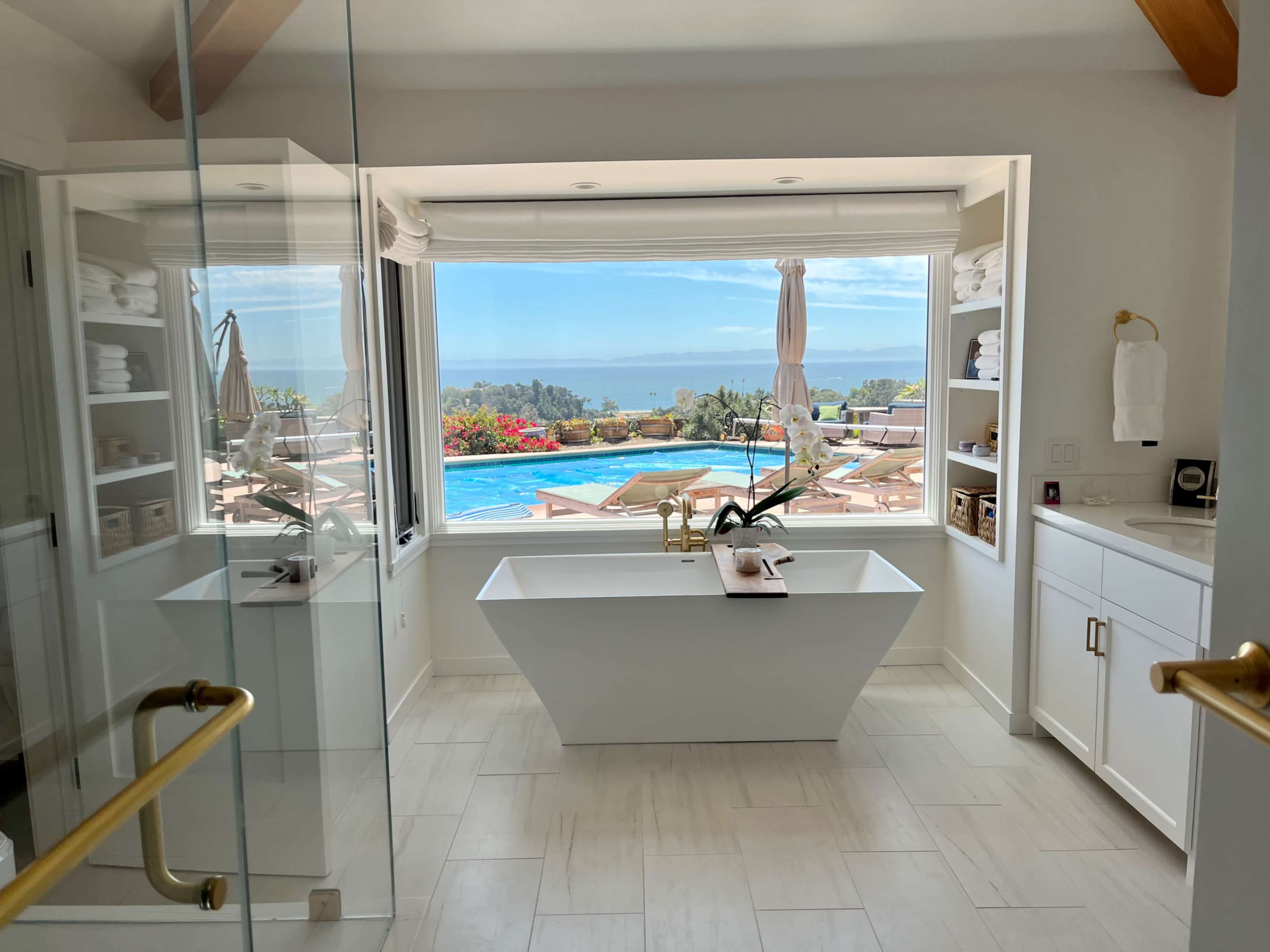 A modern bathroom with a freestanding bathtub and a large window overlooking a pool and ocean view.