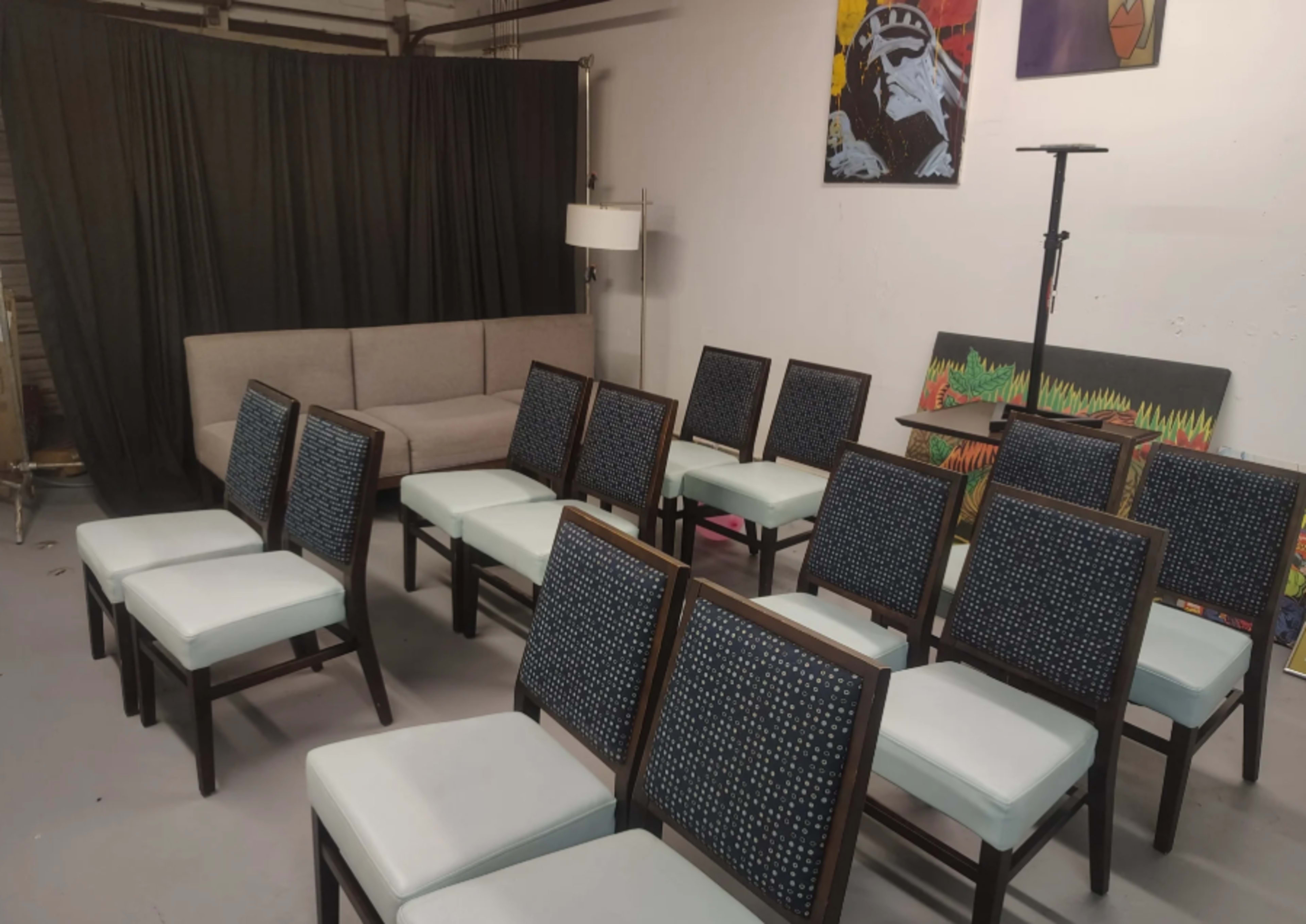 The image shows a room with a gray sofa against a black curtain, surrounded by several wooden chairs with light-colored cushions arranged in rows.