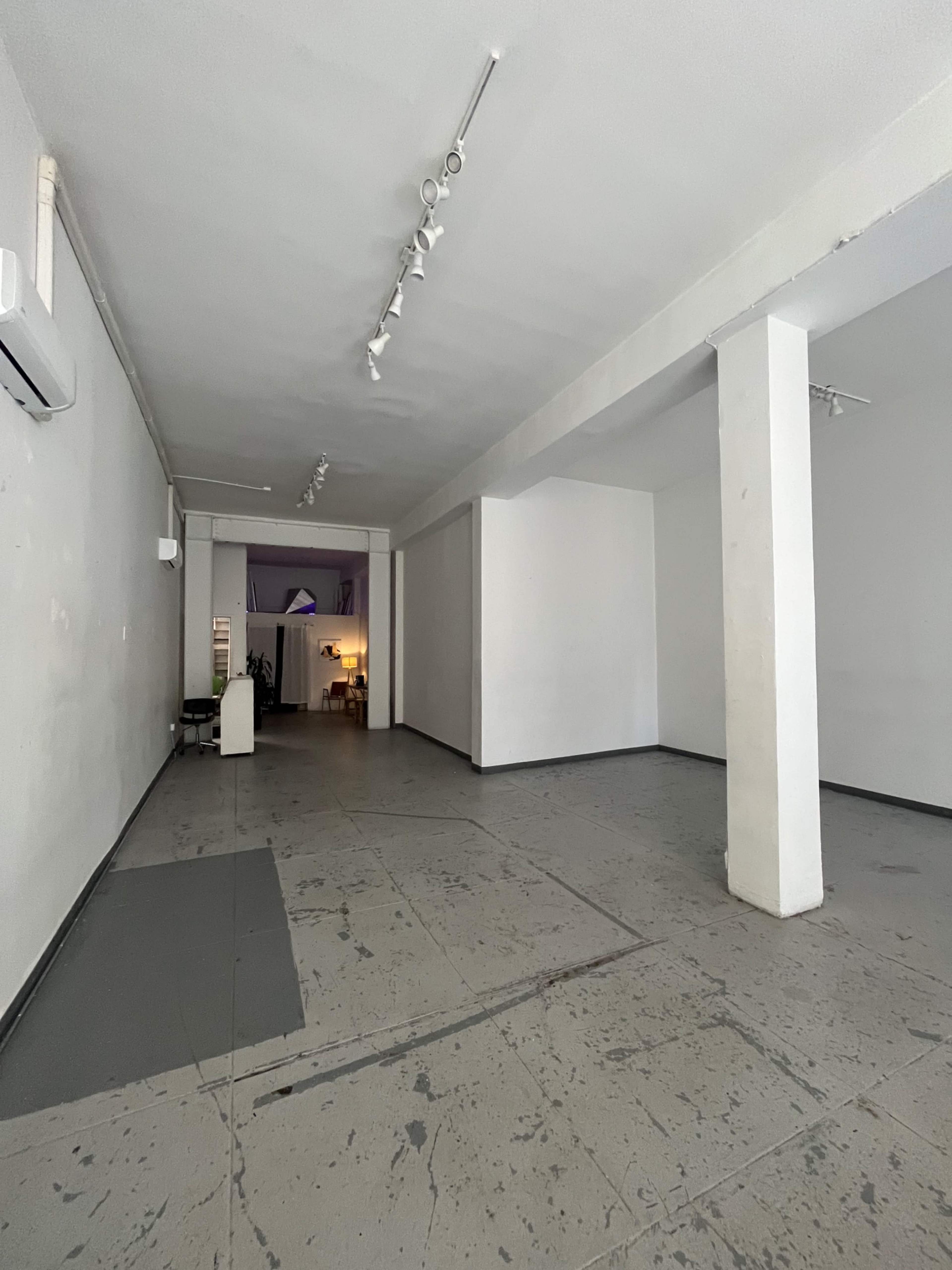 An empty, minimalist interior space with gray floors and white walls, featuring several columns and minimal furniture visible in the background.