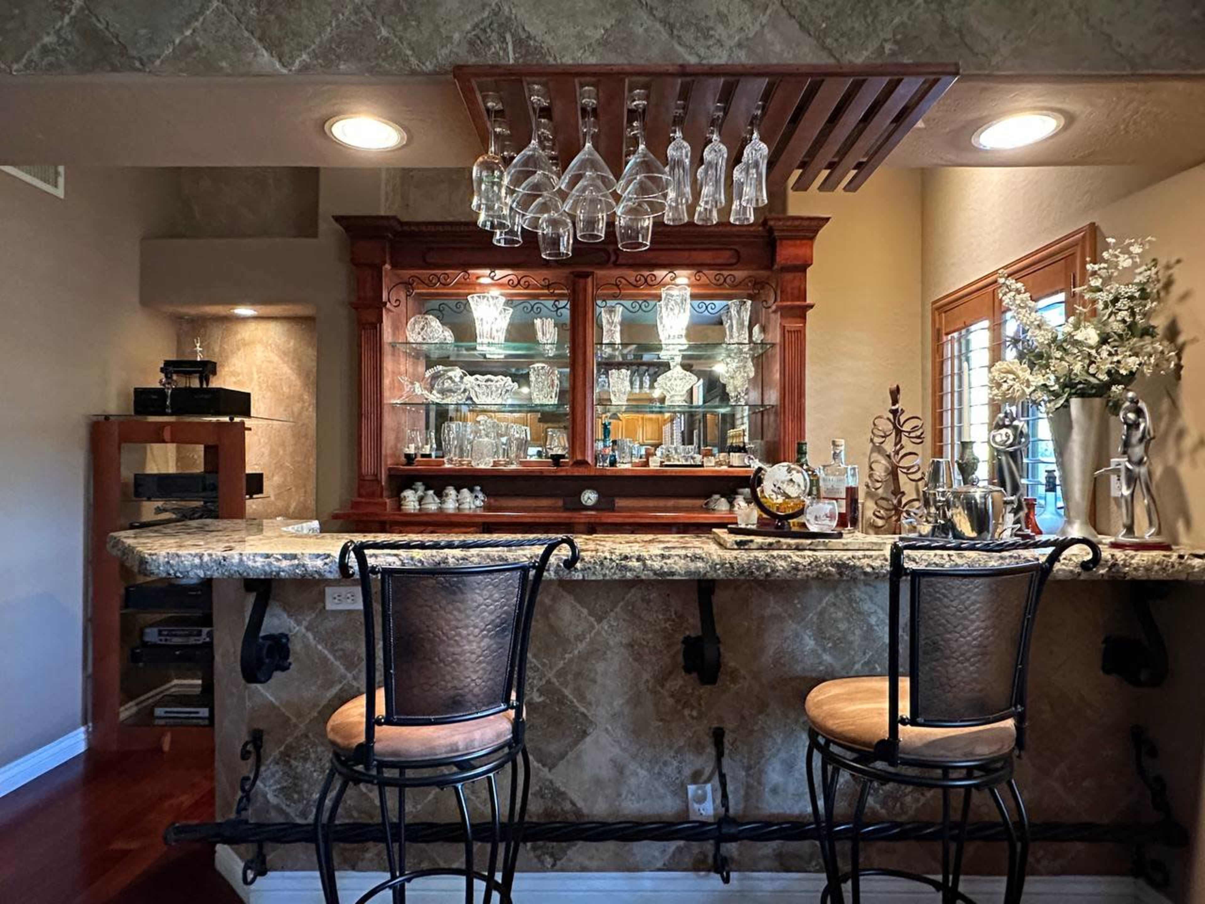 A home bar area features a granite countertop, wooden cabinetry with glass displays, and two high-backed stools.