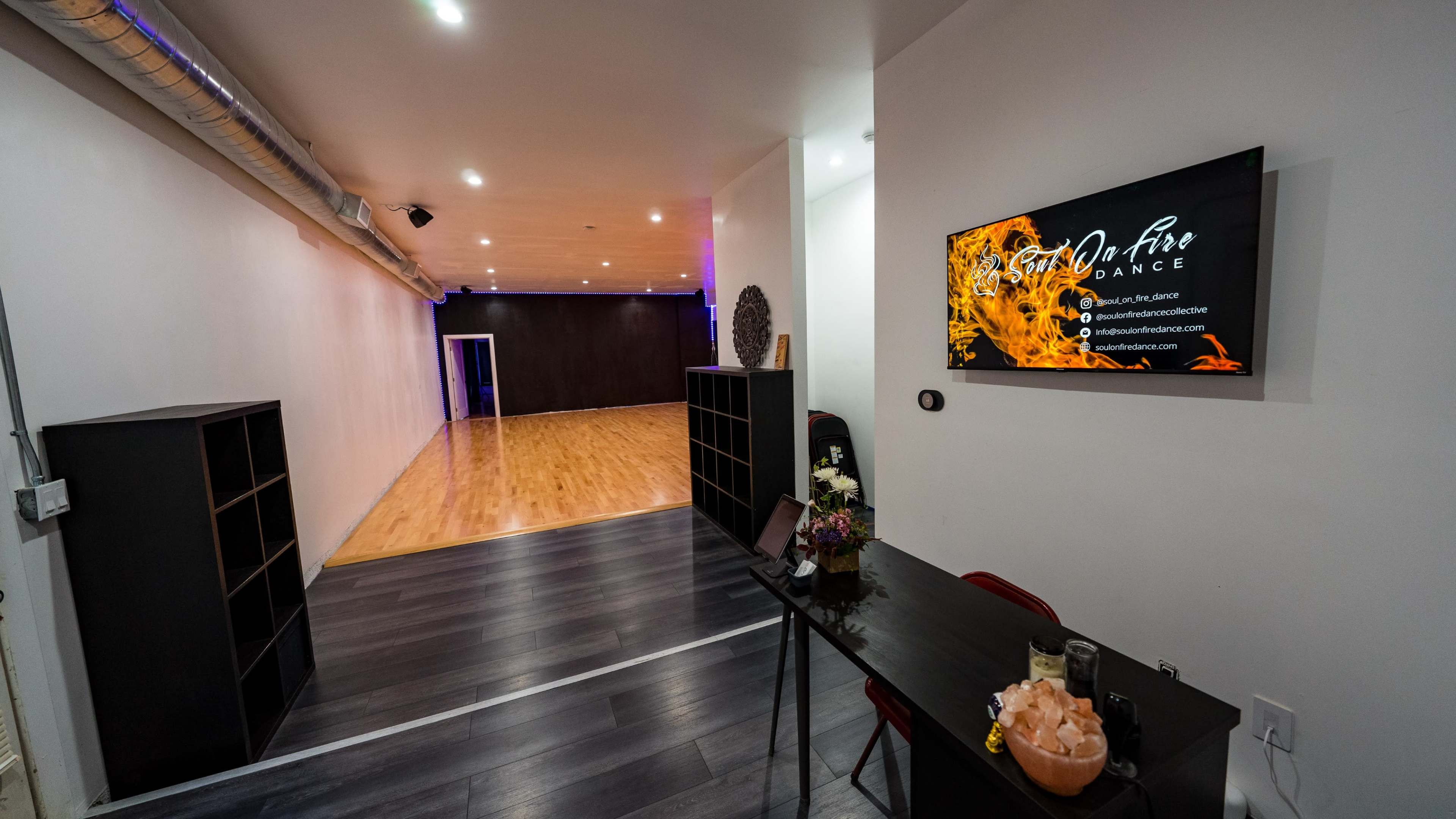 A spacious dance studio with wooden flooring, a reception area featuring a small table and flowers, and a screen displaying the name of the studio on the wall.