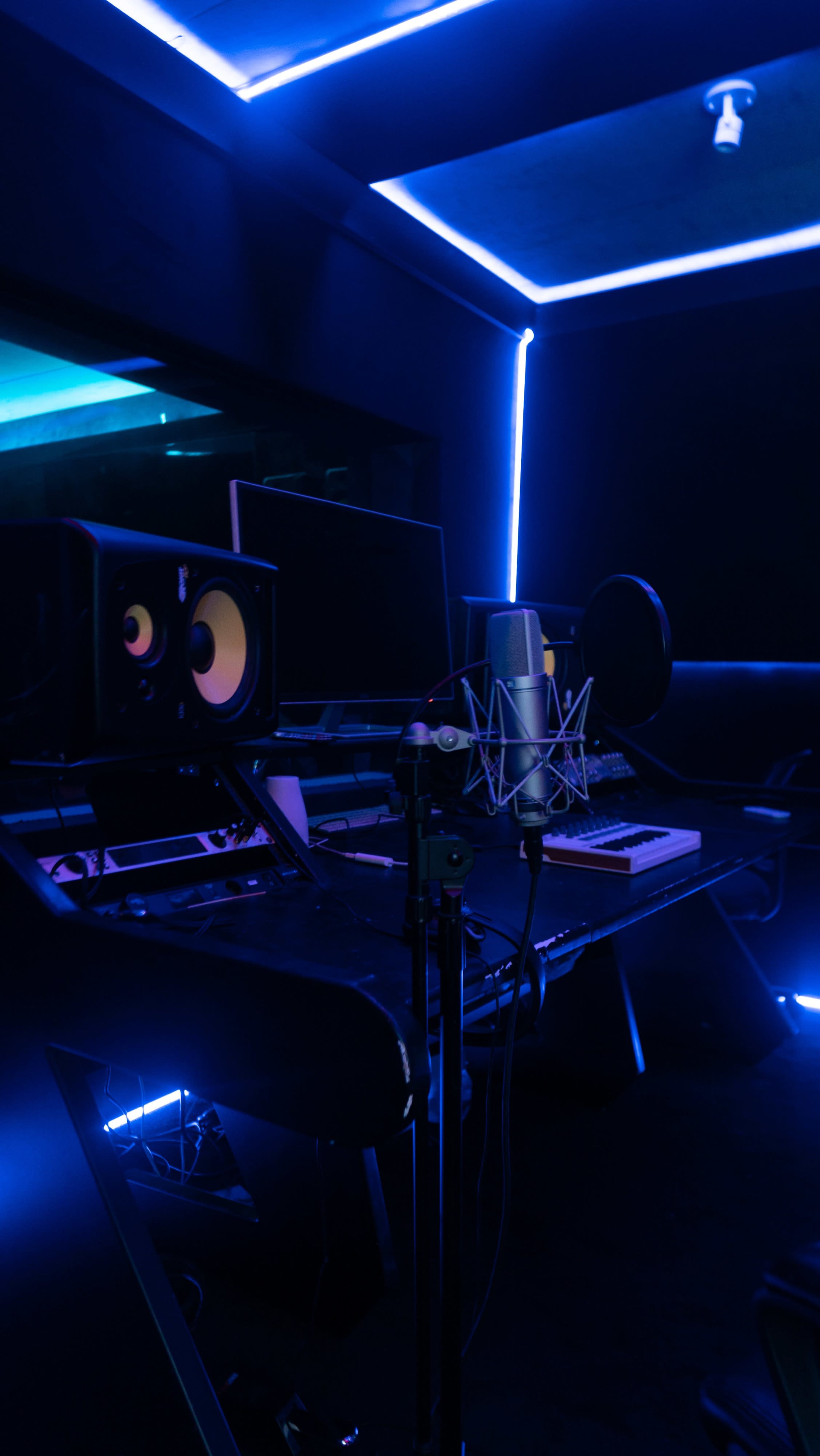 A music recording studio features a microphone, computer monitors, and audio speakers illuminated by blue LED lights.