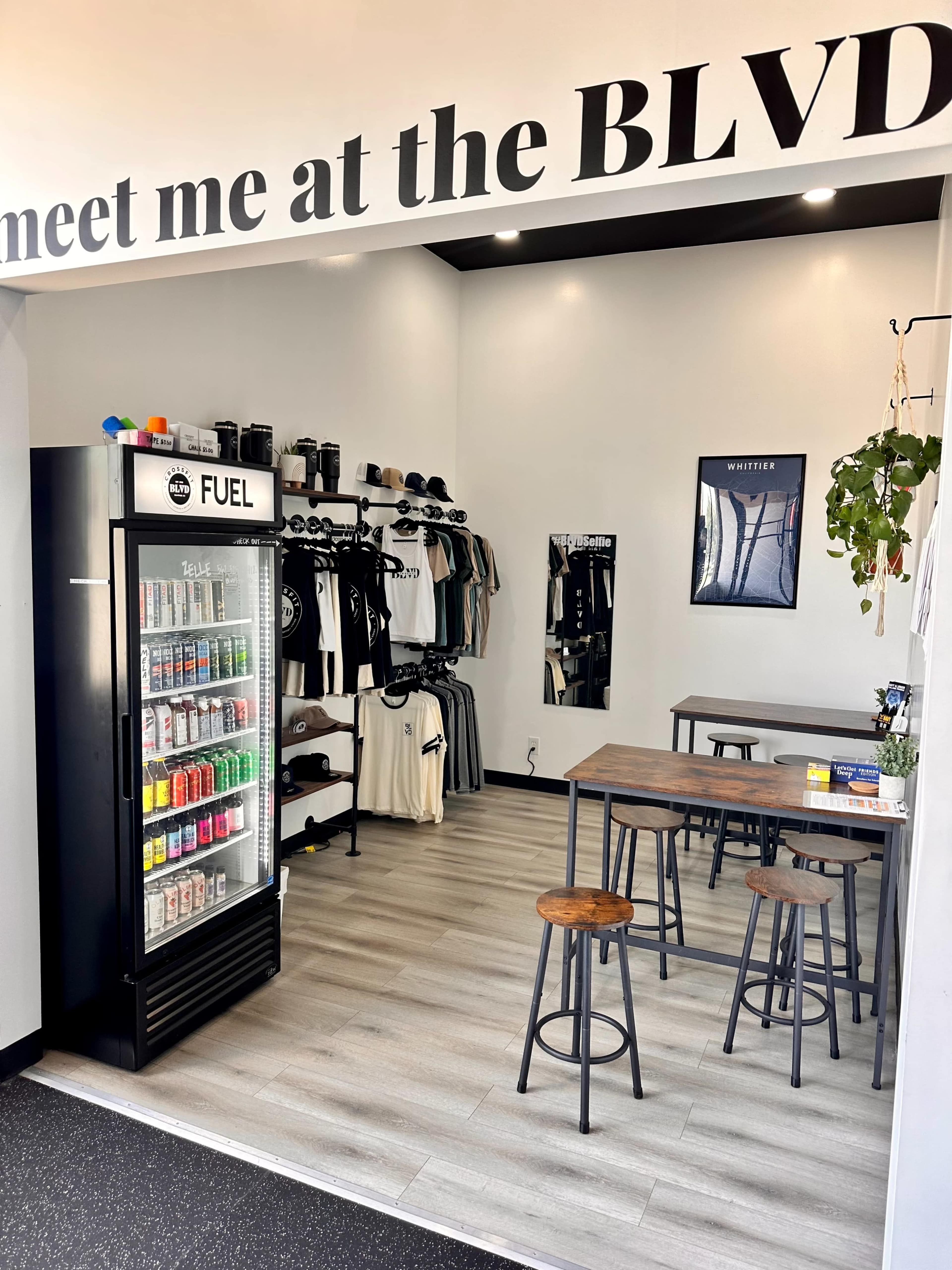 A small retail space featuring a beverage fridge labeled "FUEL," a rack of clothing on hangers, and two wooden tables with stools.