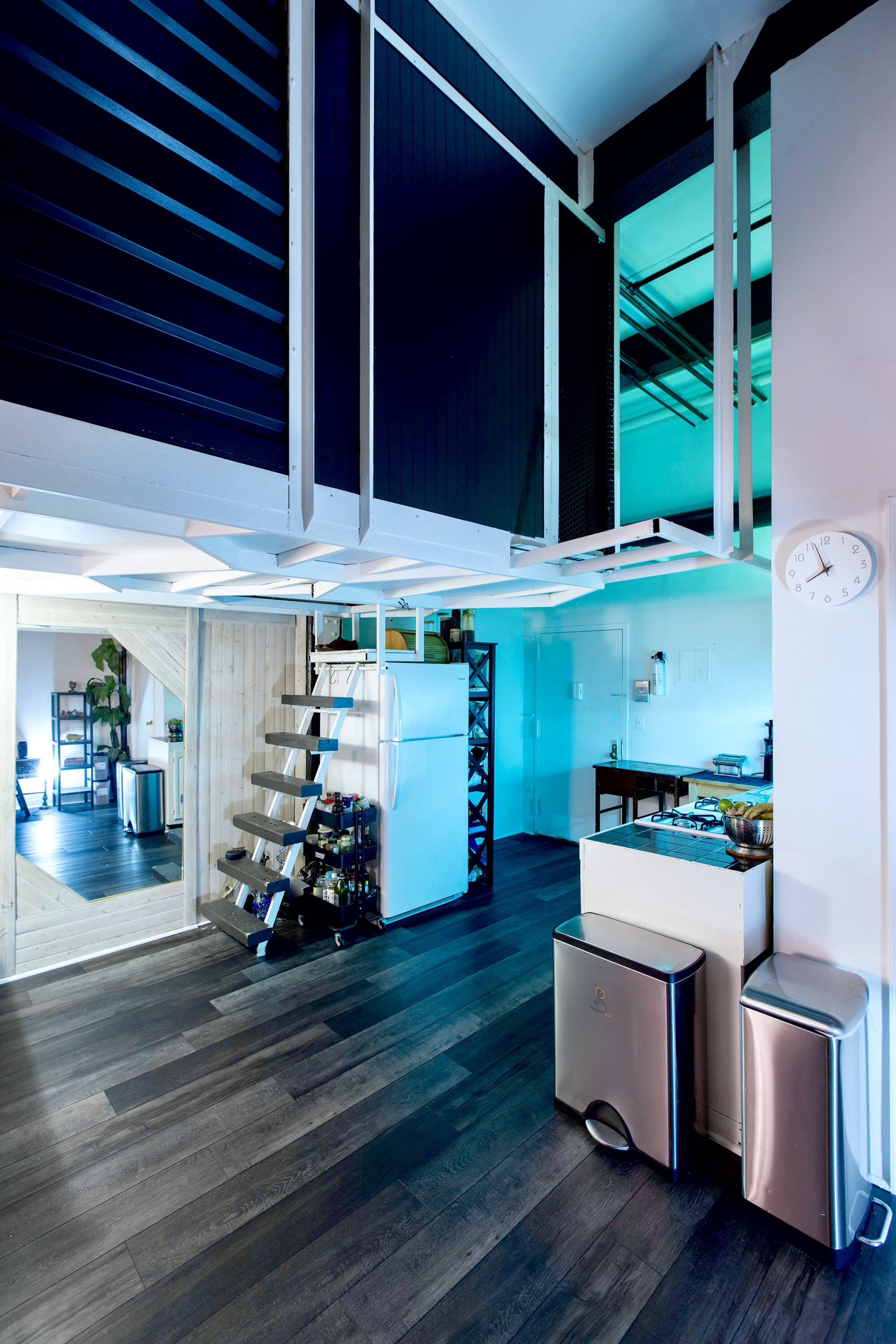 The image shows a modern, open-space interior with a staircase leading to a lofted area, a kitchen featuring appliances, and a large mirror reflecting the space.