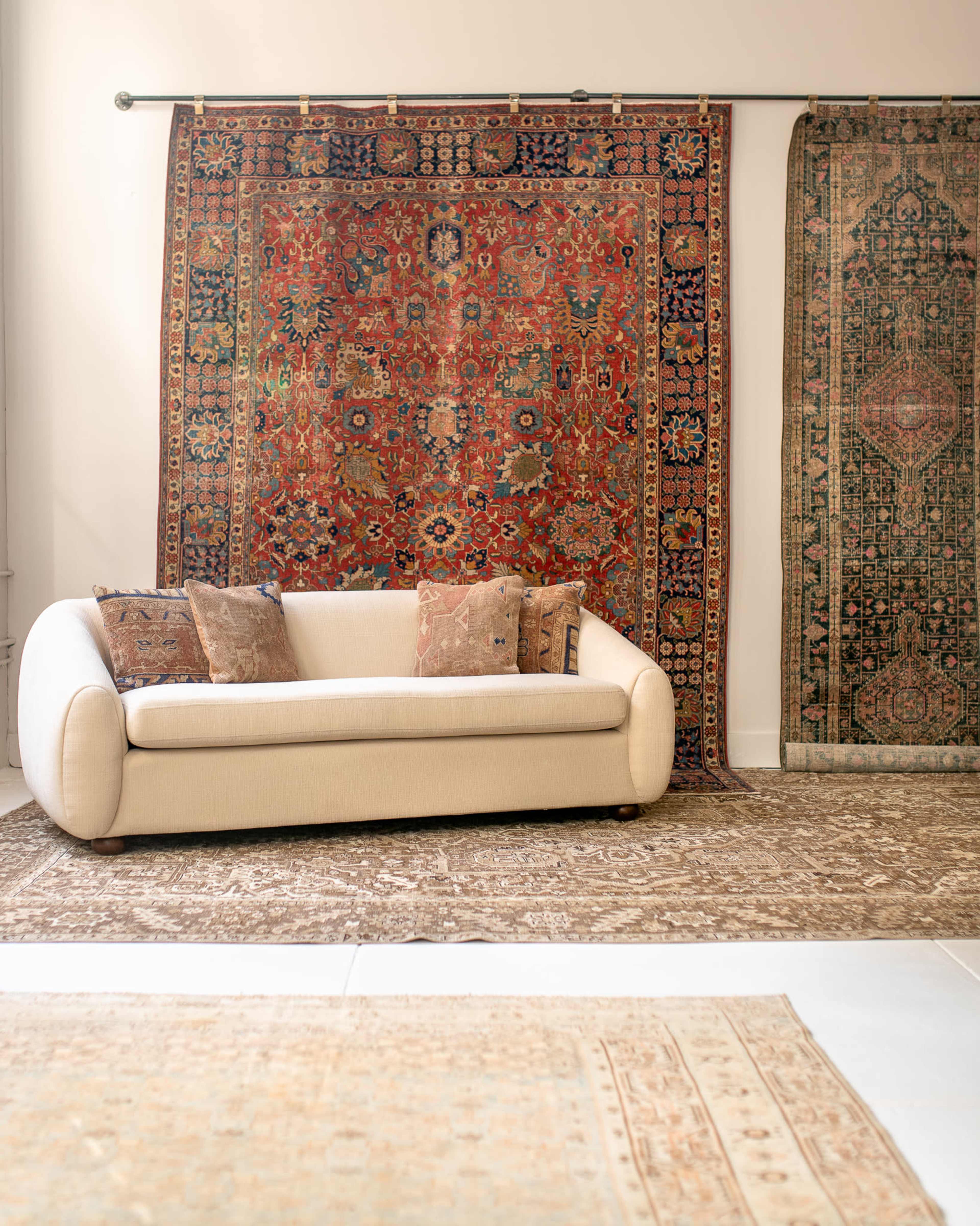 A beige sofa is positioned in front of two large hanging carpets on a light-colored wall, with smaller decorative pillows placed on the sofa.
