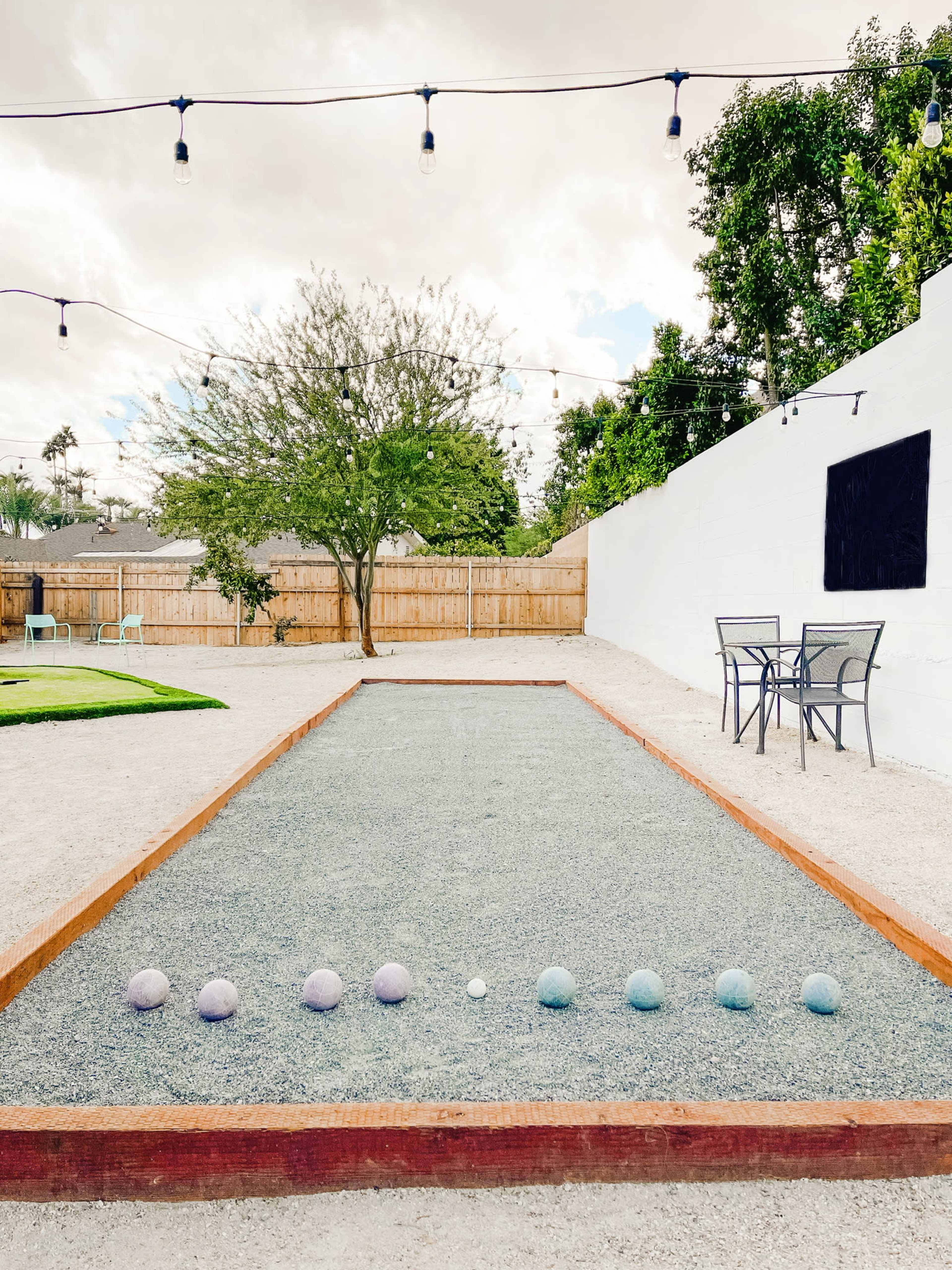 A gravel bocce court with colorful balls lined up, surrounded by greenery and string lights, is set against a white wall and a wooden fence.