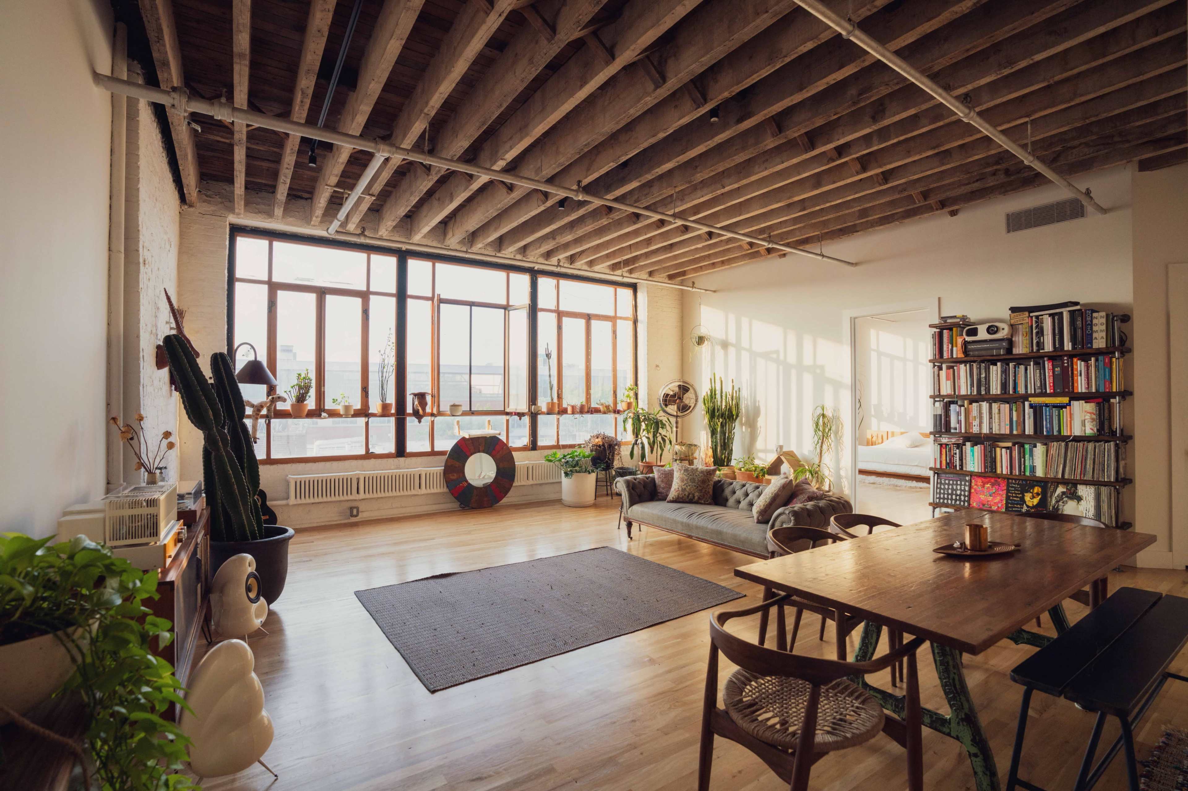 The image shows a spacious loft-style living area with large windows, a wooden ceiling, and rustic furnishings, including a sofa, dining table, and various plants.