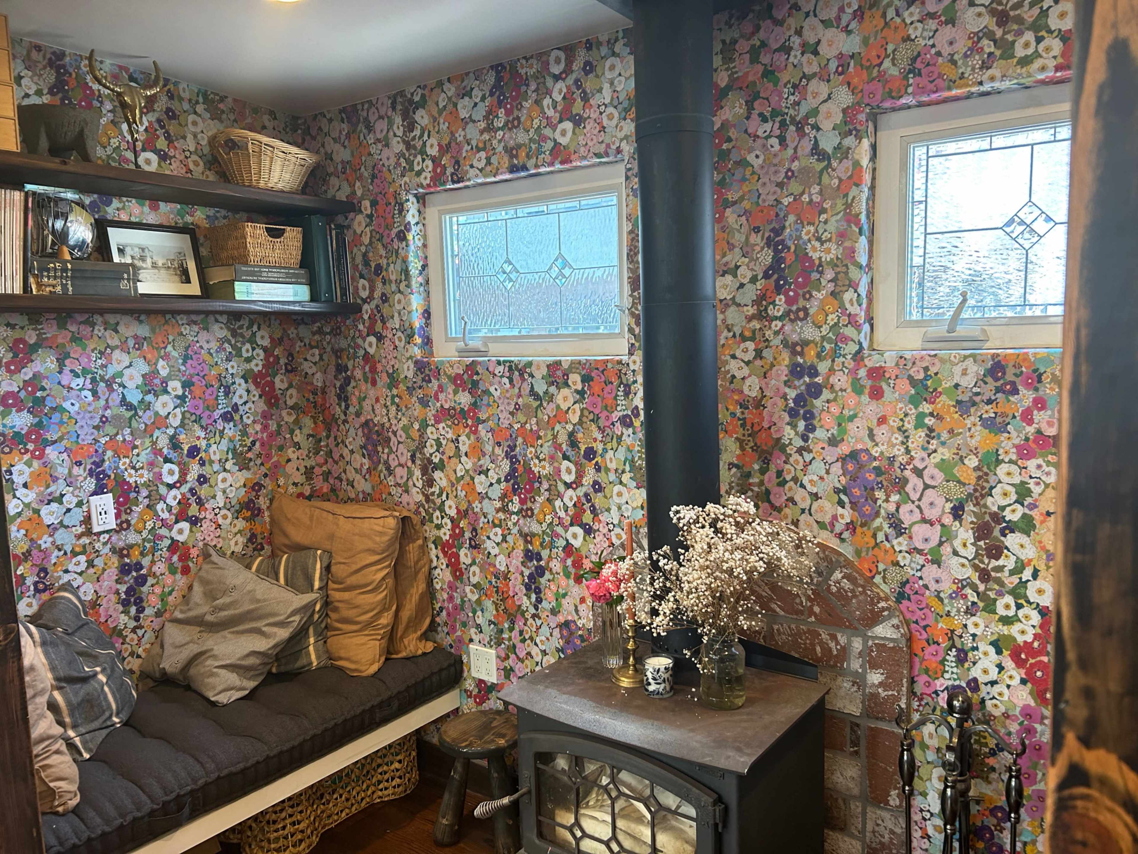 The image shows a cozy corner of a room with flower-patterned walls, a seating area with cushions, a small table with a decorative vase, and a wood-burning stove.