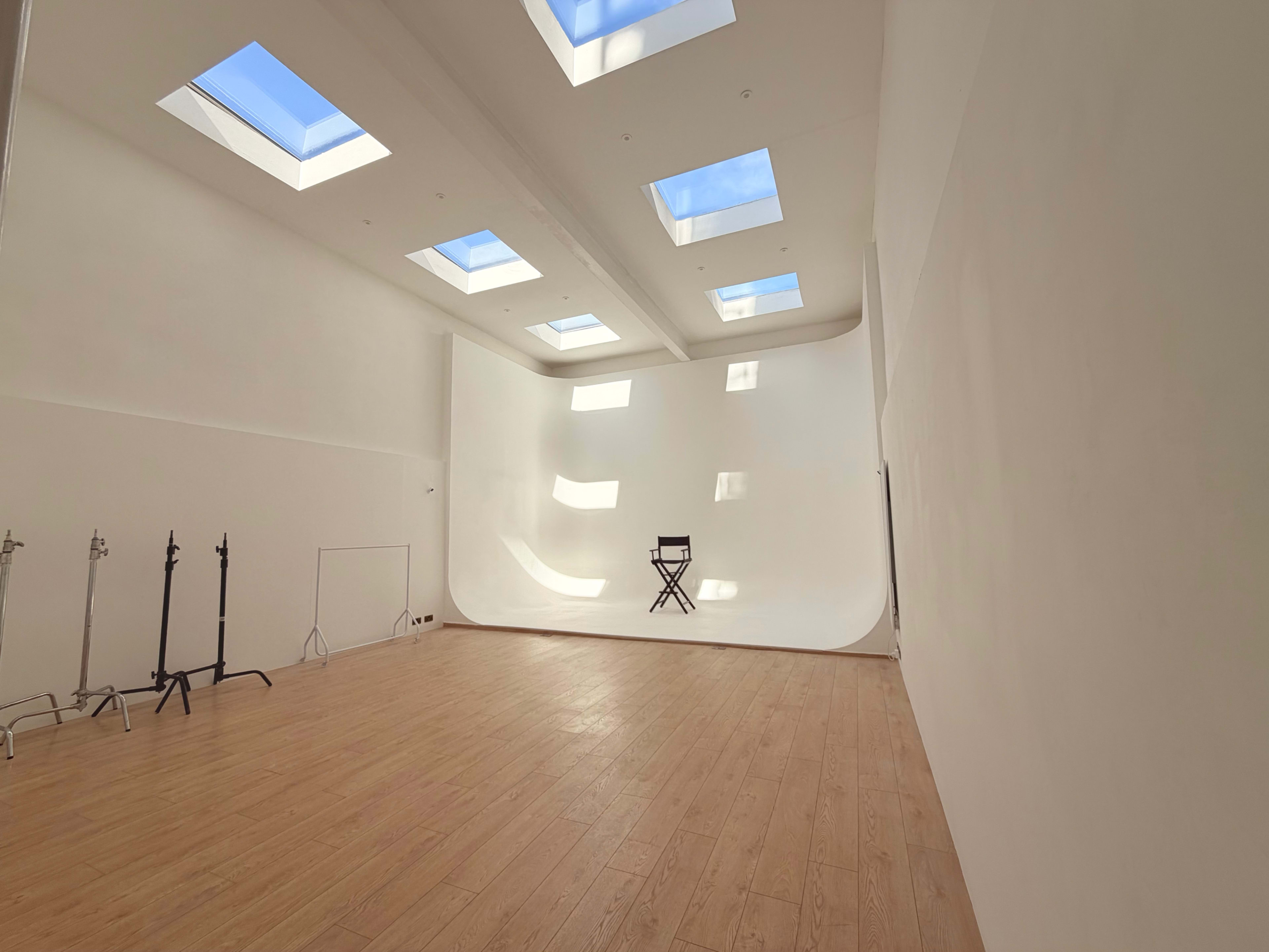 The image shows a spacious photography studio with a curved white backdrop, wooden flooring, and several skylights allowing natural light to enter.