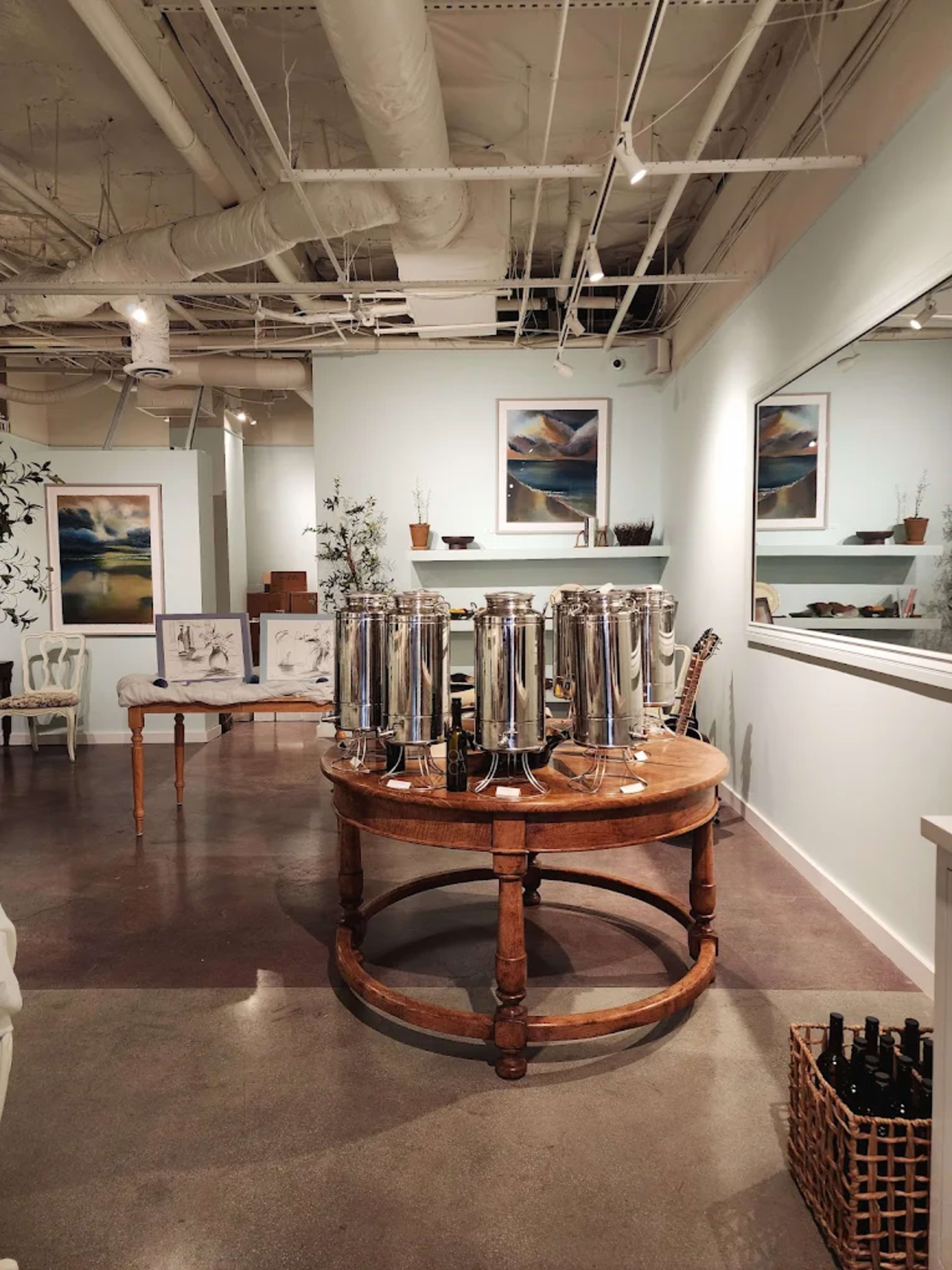The image shows a spacious room with a round wooden table at the center, featuring several shiny metal beverage dispensers, surrounded by framed artworks and decor elements.