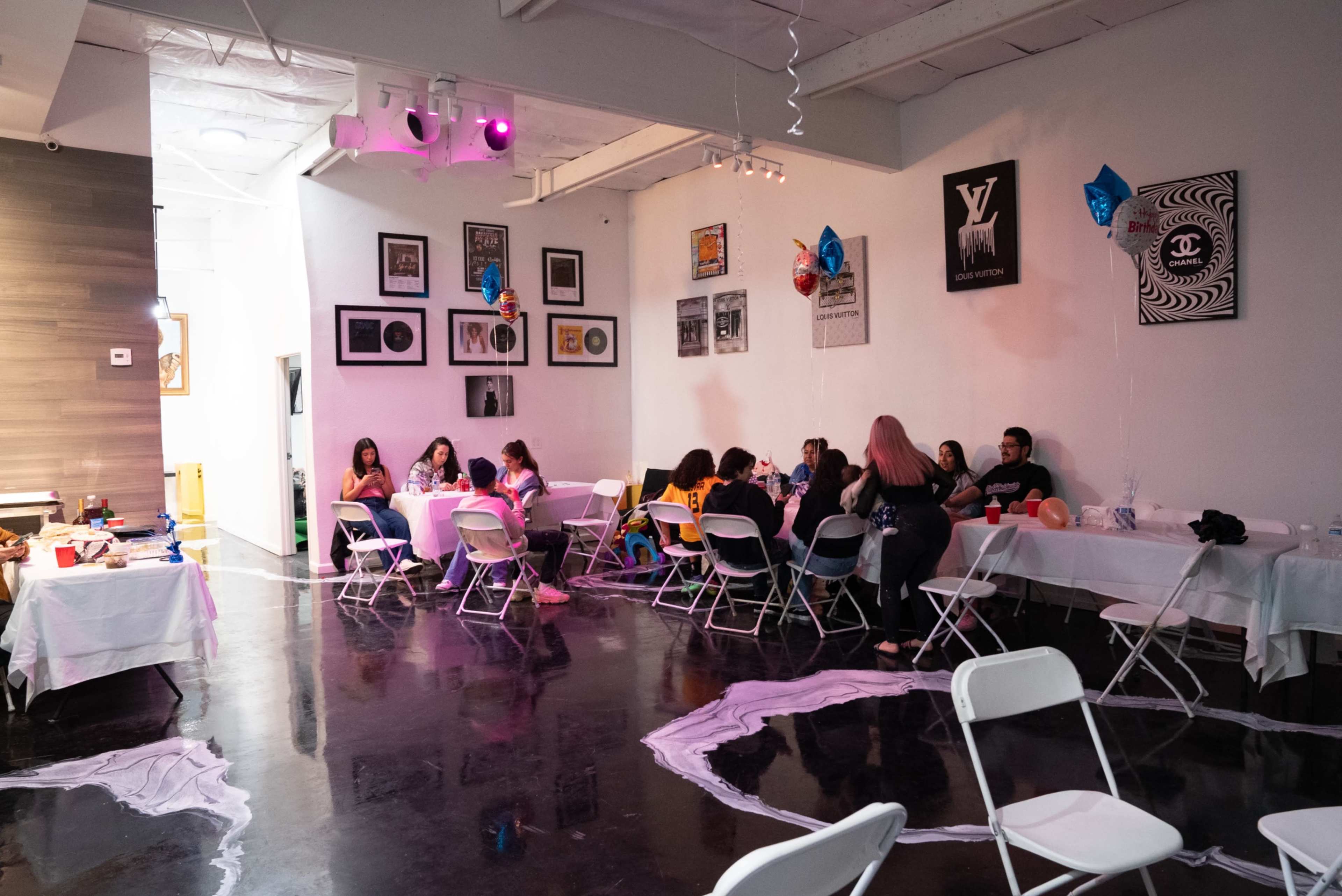 A group of people gathers around tables in a modern room decorated with balloons and artwork, while white tablecloths cover the surfaces.