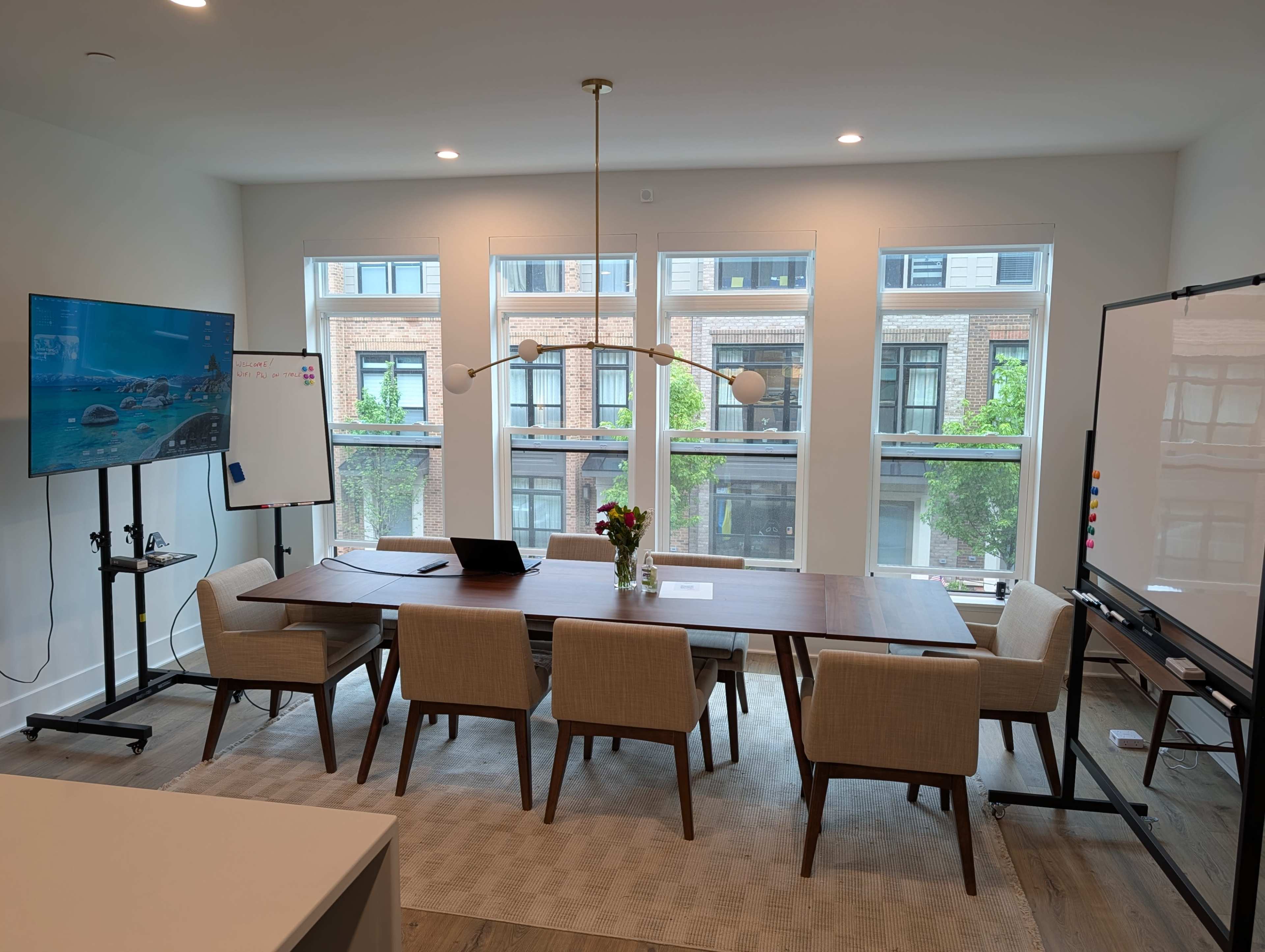 A modern conference room features a long wooden table with chairs, a large screen on a stand, a whiteboard, and ample natural light from large windows.