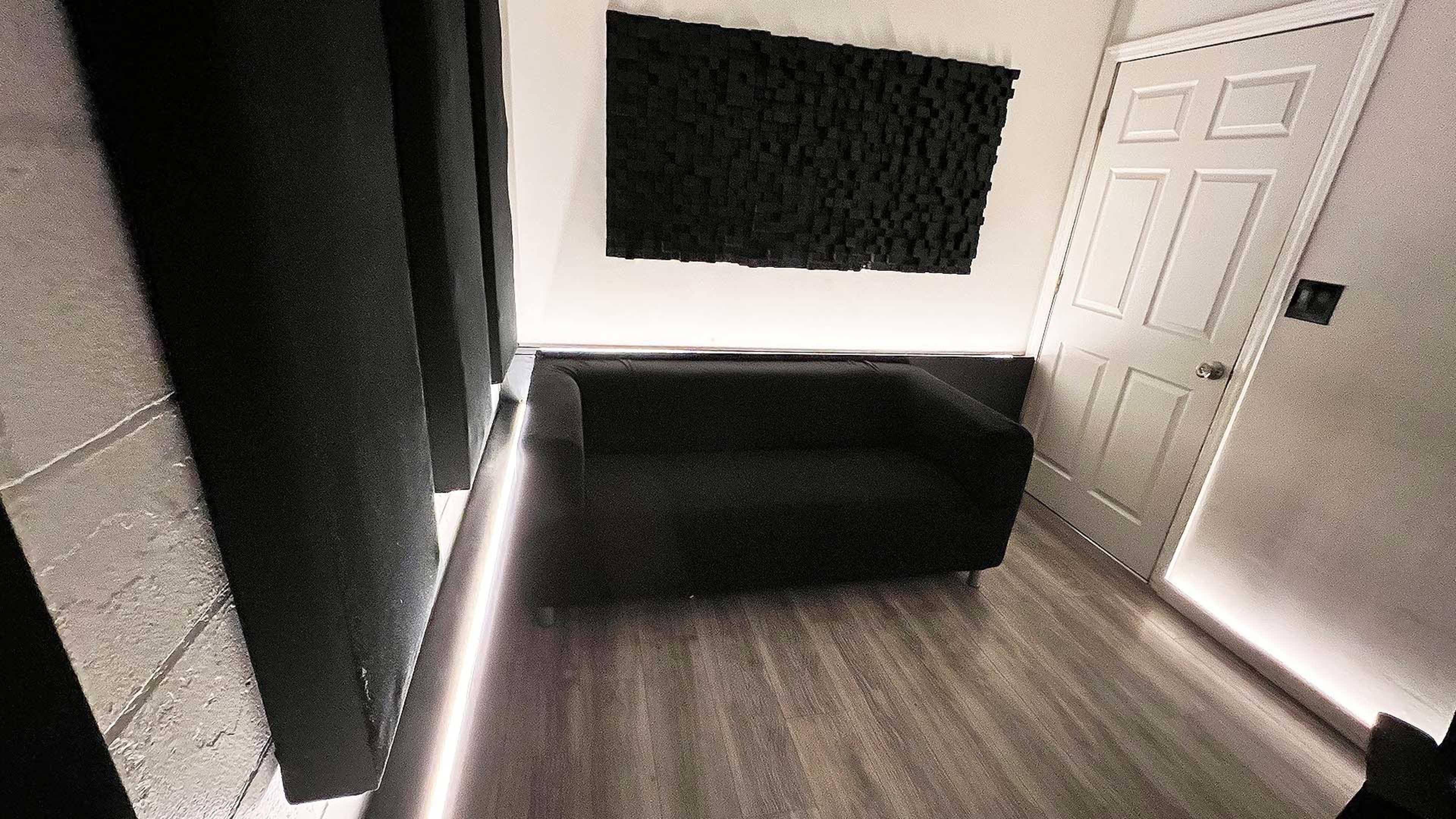 A modern room with a black couch, acoustically treated wall panels, and dim LED lighting along the baseboards.