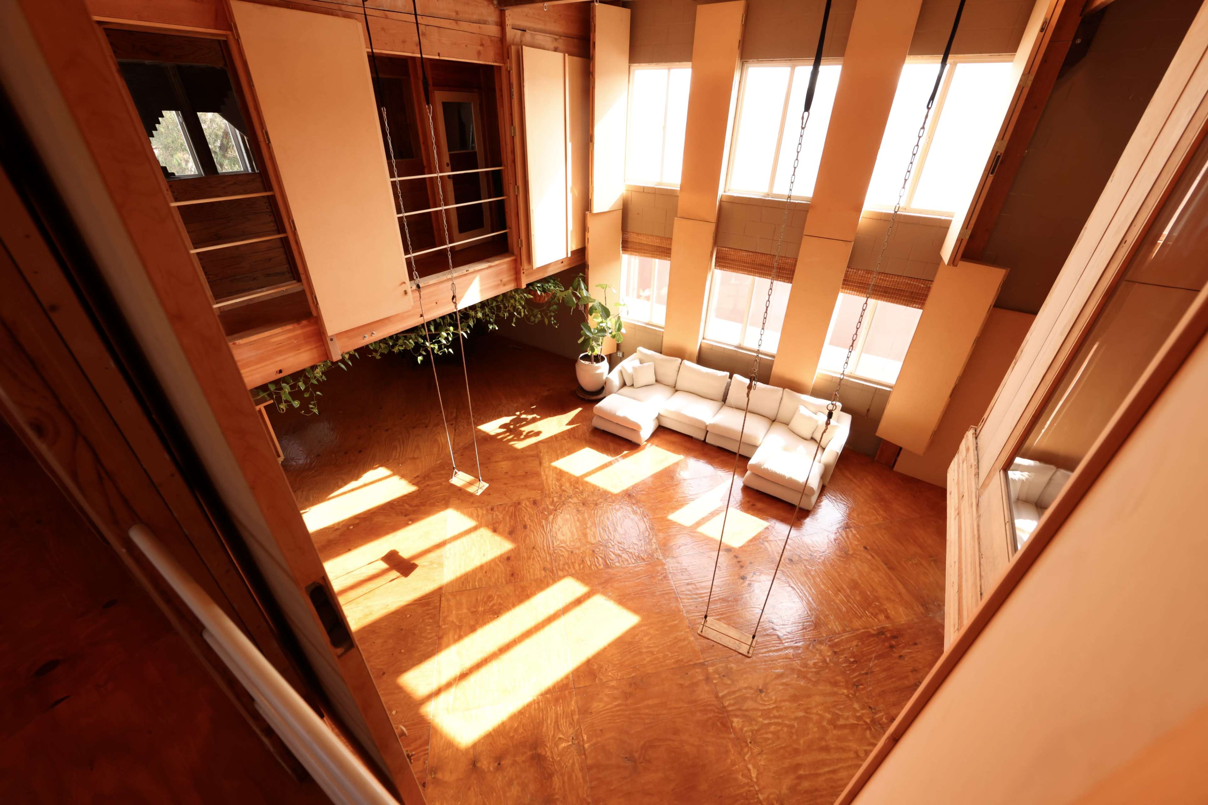 The image shows a spacious interior featuring a white sofa, large windows letting in sunlight, and a unique wooden architecture with swings hanging from the ceiling.