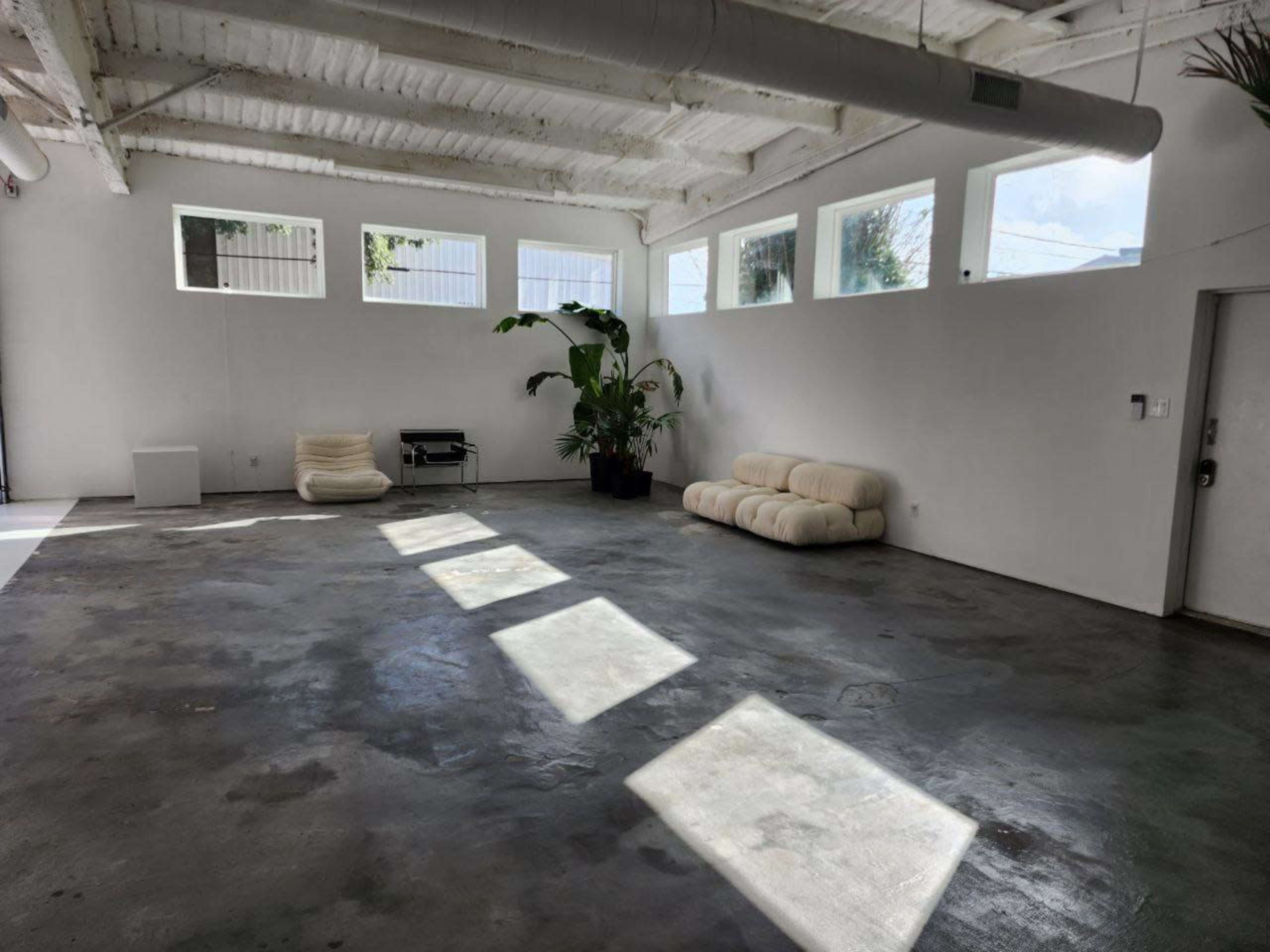 The image shows a minimalist room with concrete floors, large windows allowing natural light, and a couple of seating options, including a plush sofa and a bean bag.
