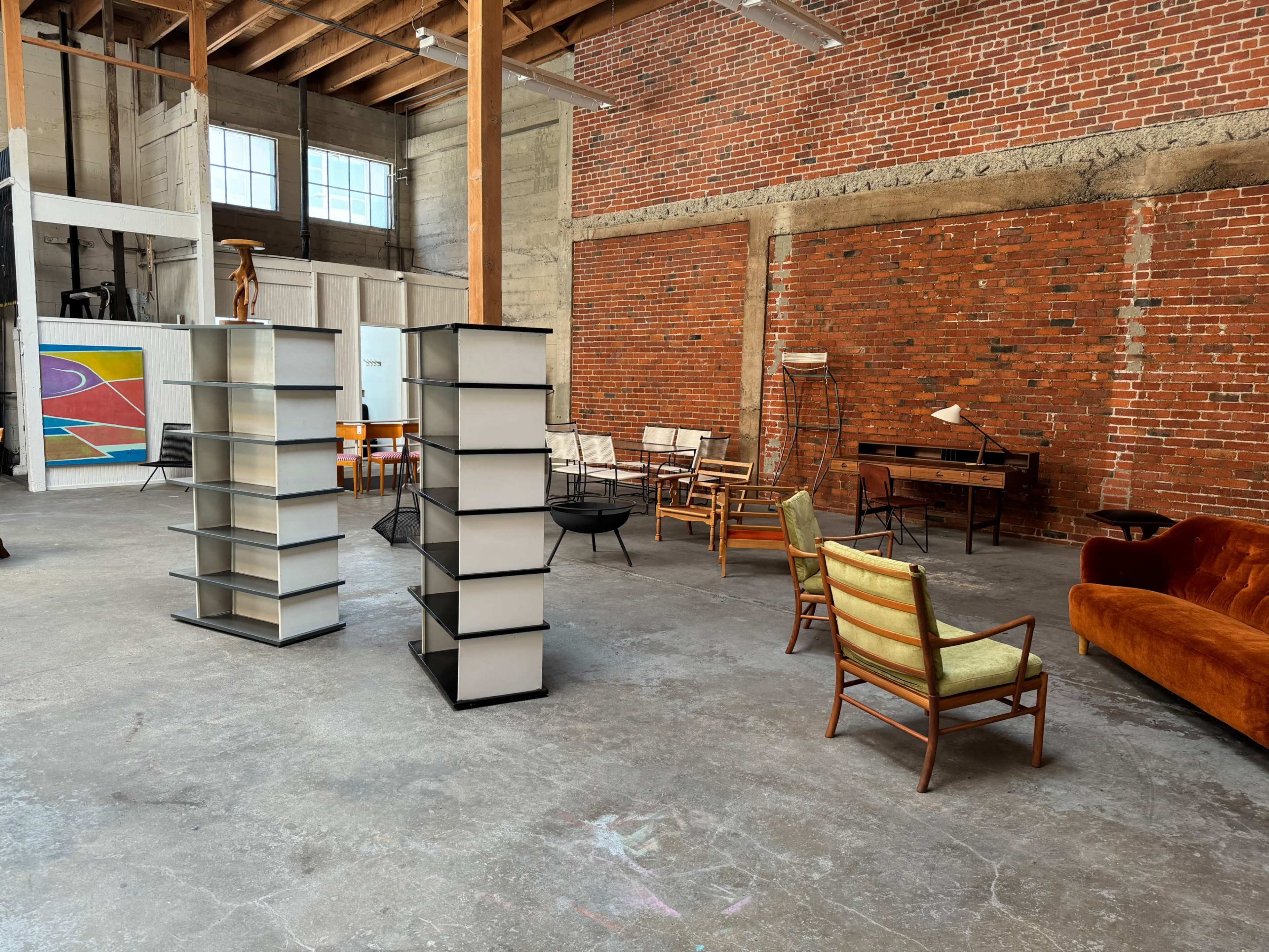 An open, industrial-style interior featuring shelving units, various chairs, and a desk arranged on a concrete floor against a backdrop of exposed brick walls.
