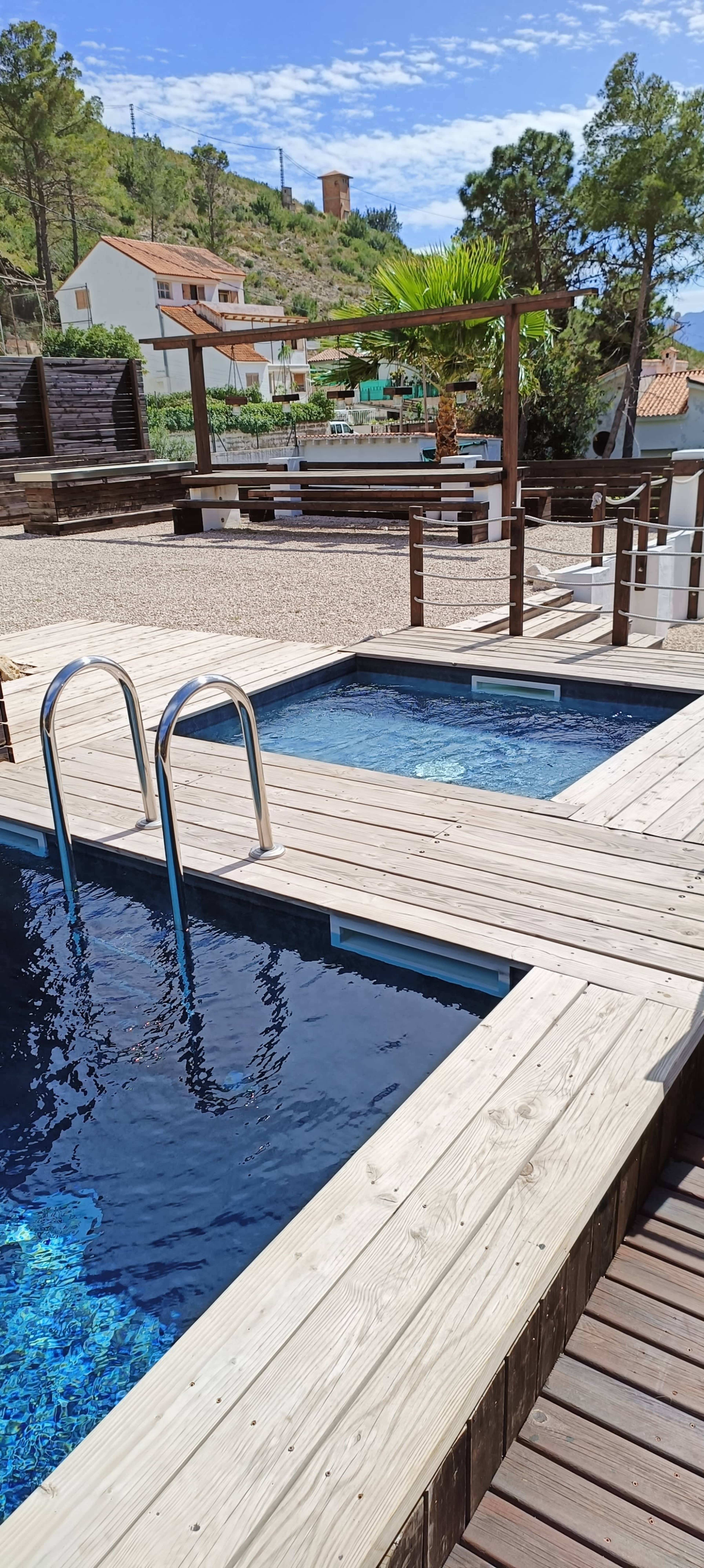 A wooden deck surrounding two swimming pools, with a landscaped area and buildings in the background.