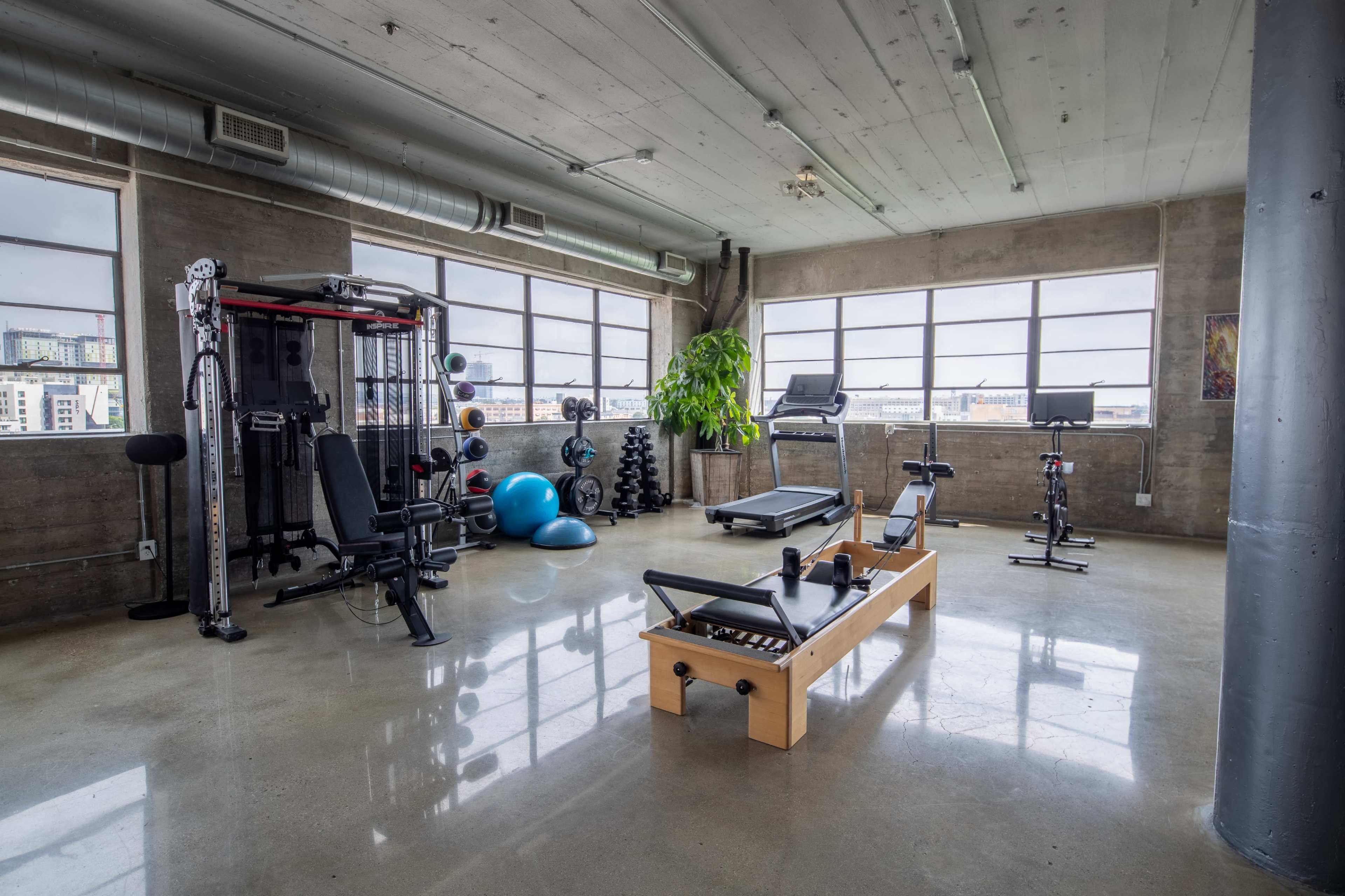 A spacious gym with various fitness equipment, including a treadmill, weight bench, free weights, and exercise balls, set against large windows.