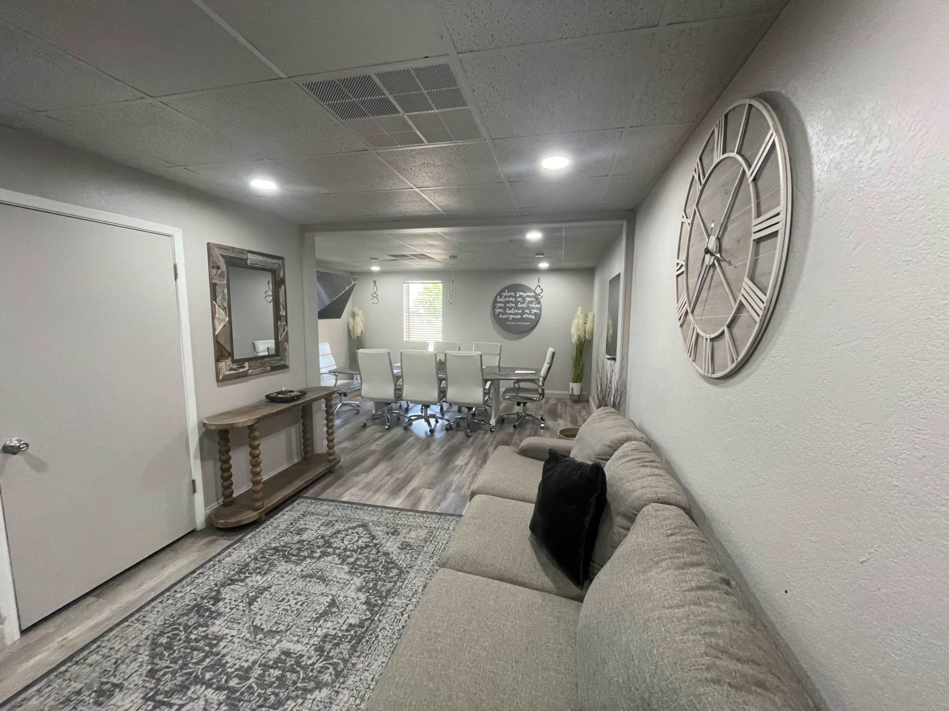 The image shows a modern office space with a seating area featuring a couch and a decorative mirror, leading to a conference room equipped with a long table and several chairs.