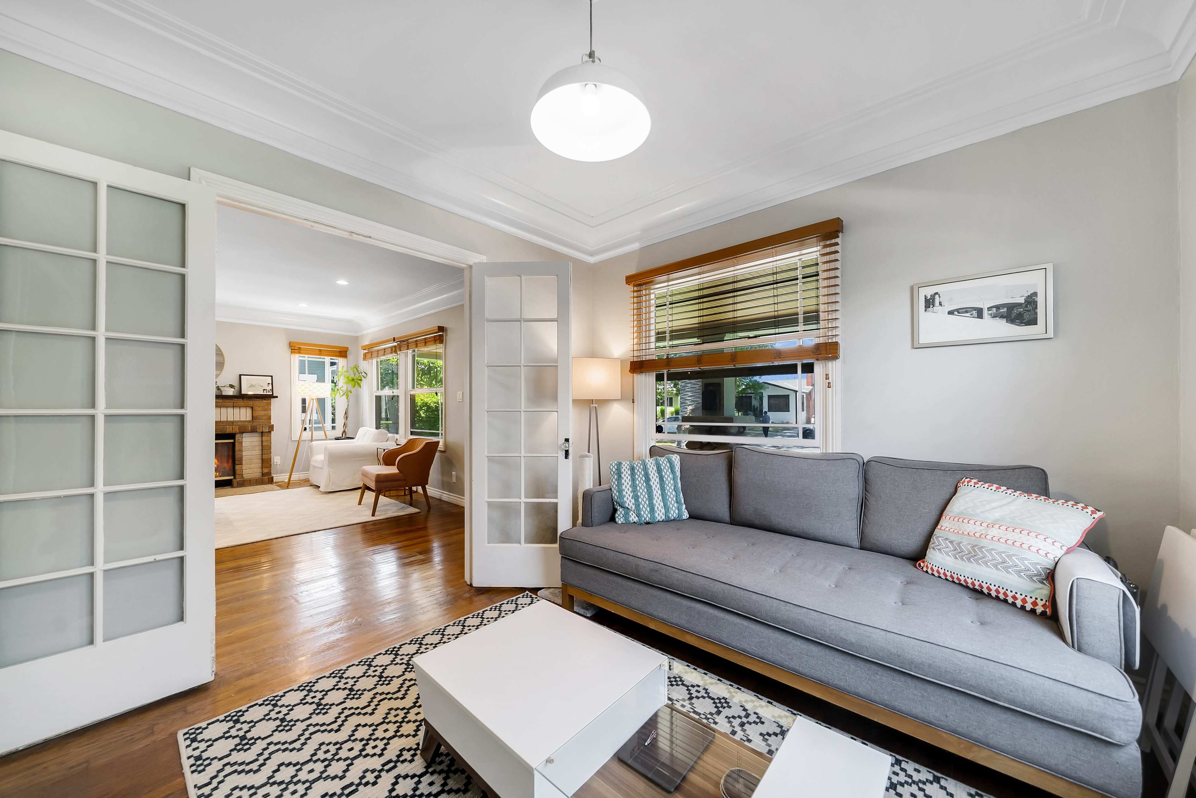 A cozy living room features a gray couch, a white coffee table, and large windows with wood blinds, leading into a bright adjacent space.