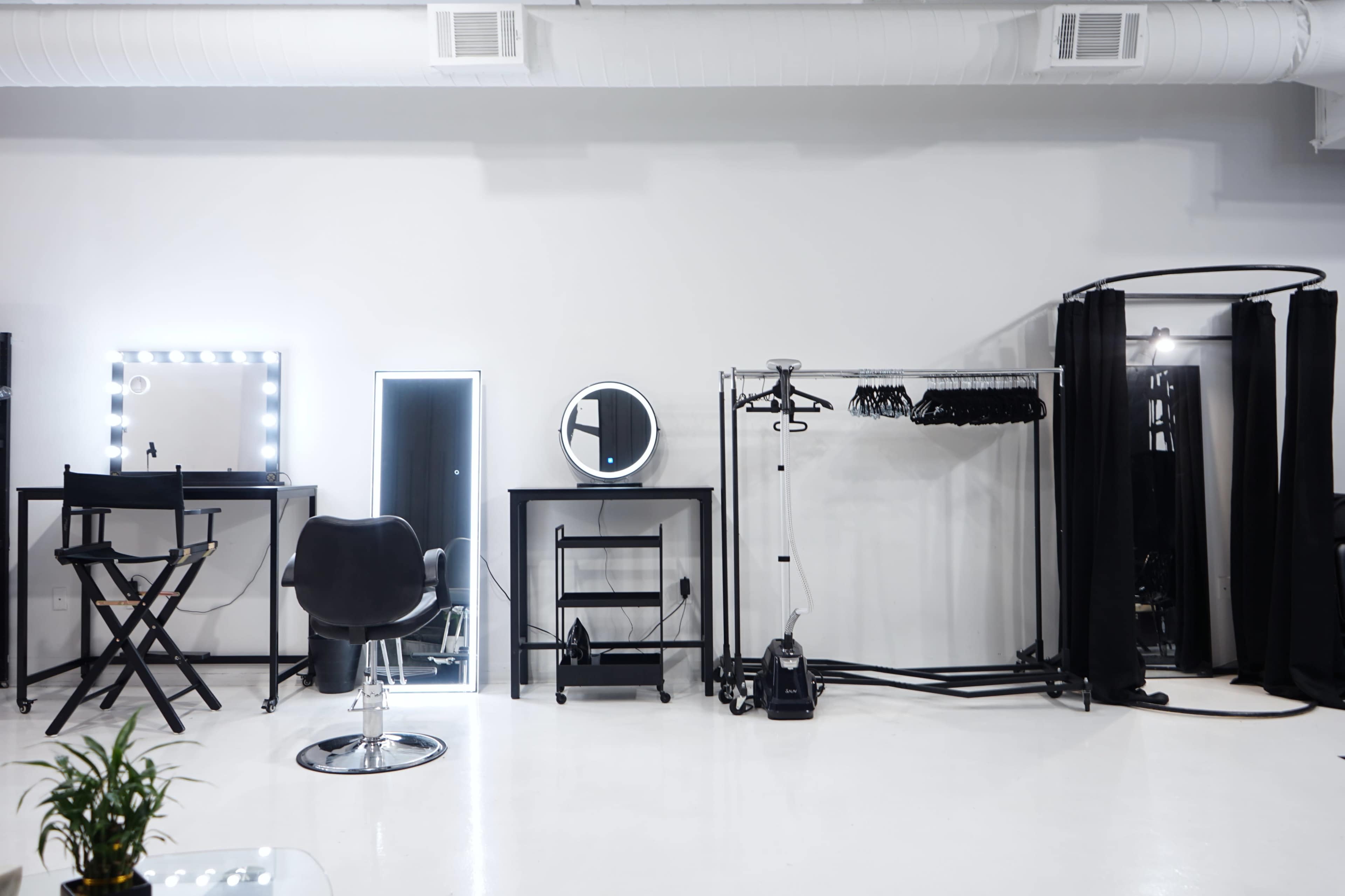 A minimalist hair salon interior featuring a styling chair, mirrors, a clothing rack, and various salon equipment against a white backdrop.