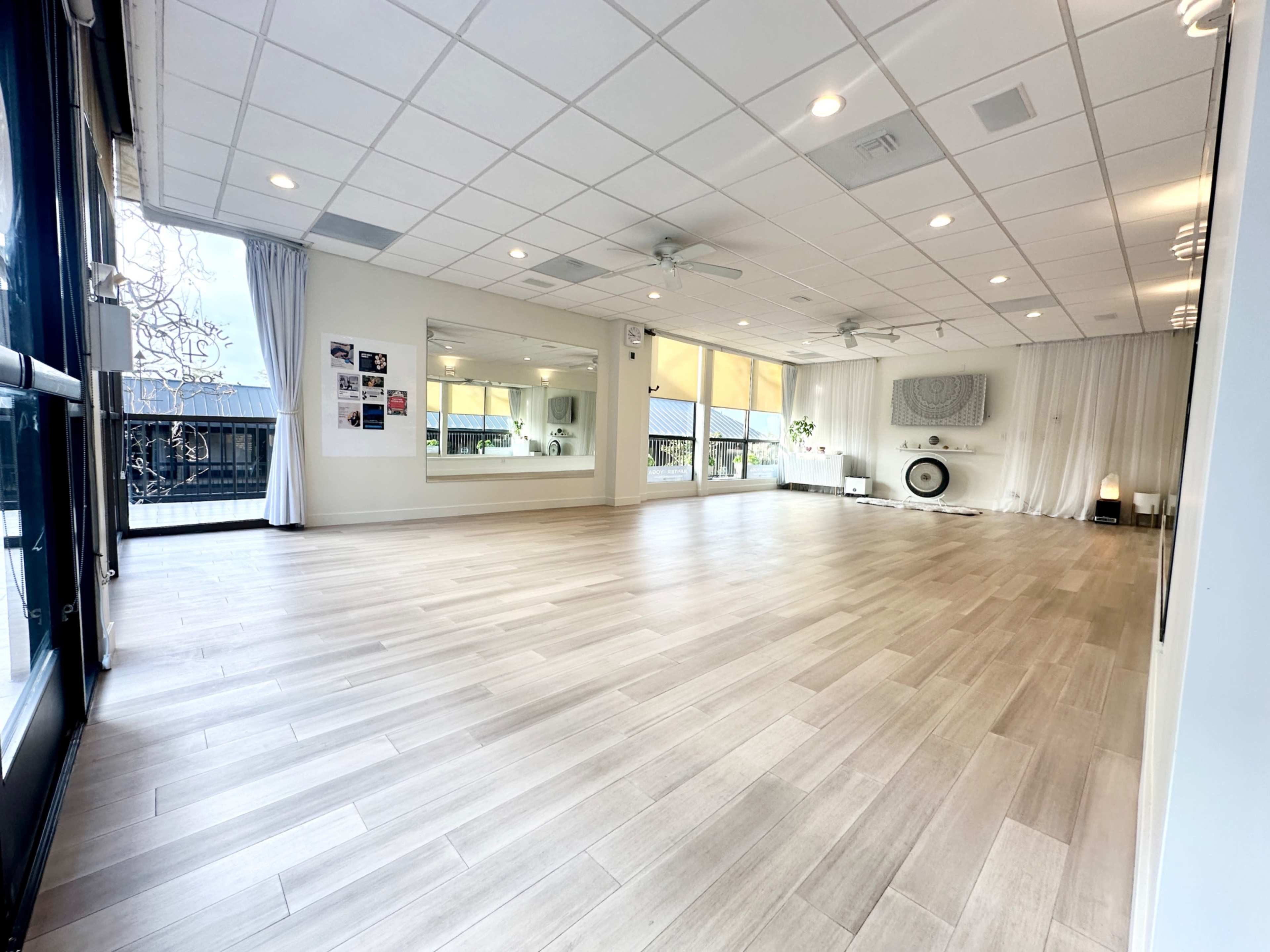The image shows a spacious, well-lit room with wooden flooring, large windows, and a minimalistic decor featuring mirrored walls and plants.