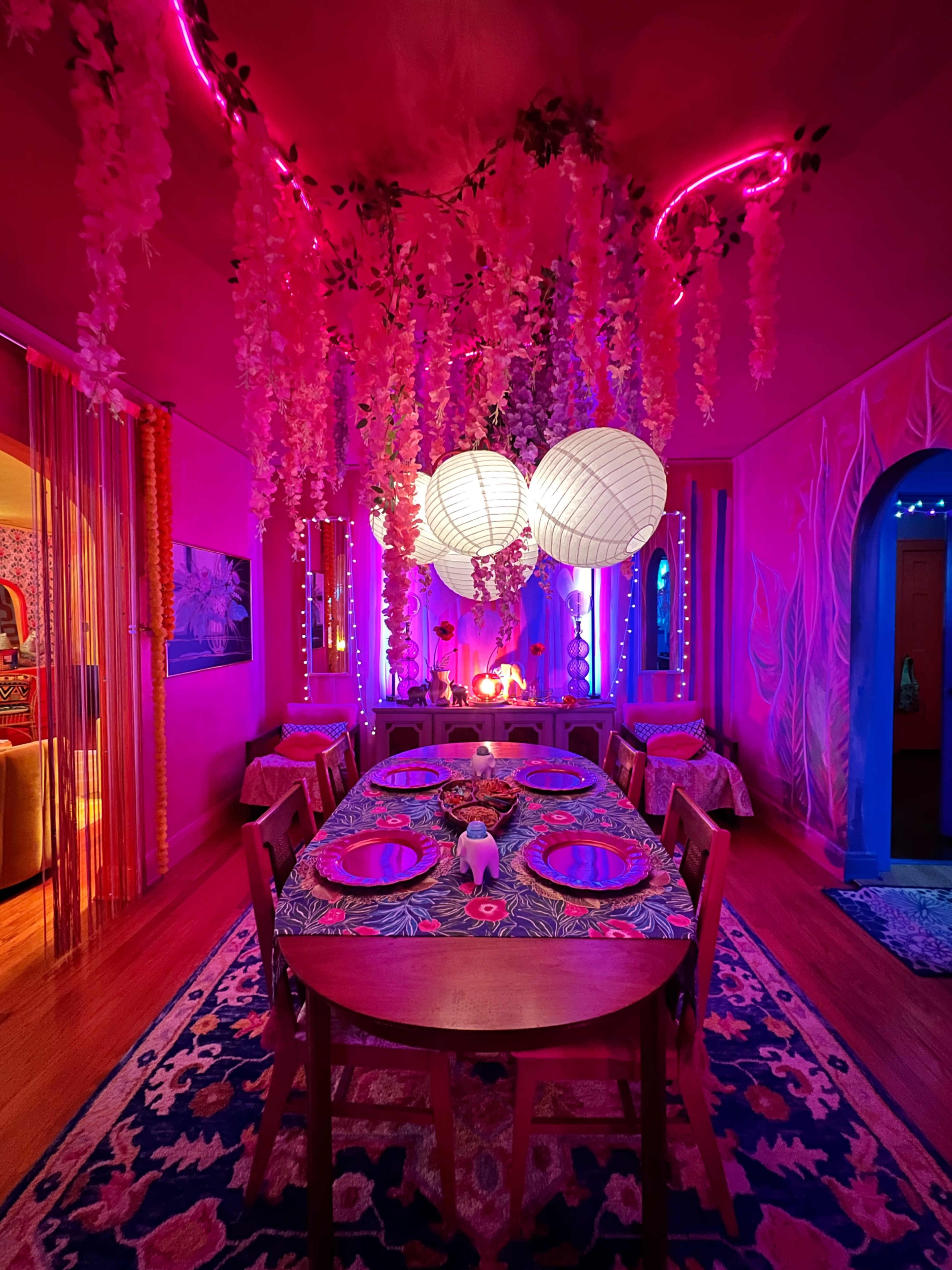 A brightly colored dining room features a long table set for a meal, adorned with decorative lighting, hanging flowers, and three large paper lanterns overhead.