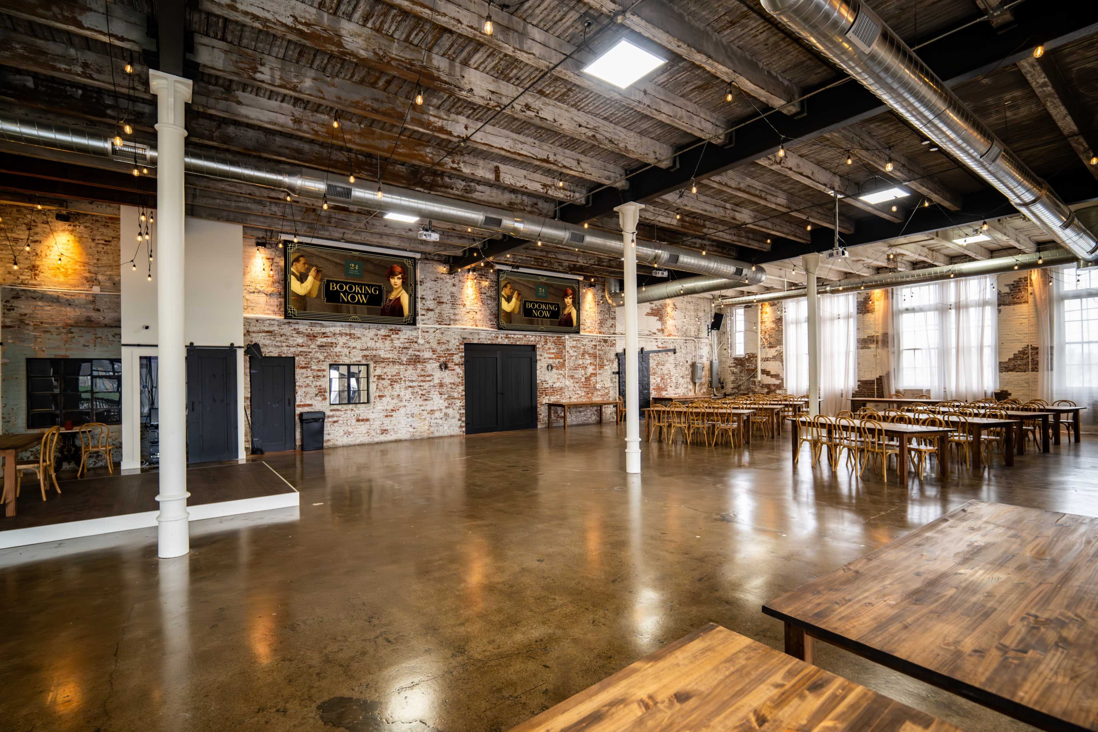 The image shows a large, industrial-style event space with exposed brick walls, polished concrete floors, and wooden tables arranged for dining.