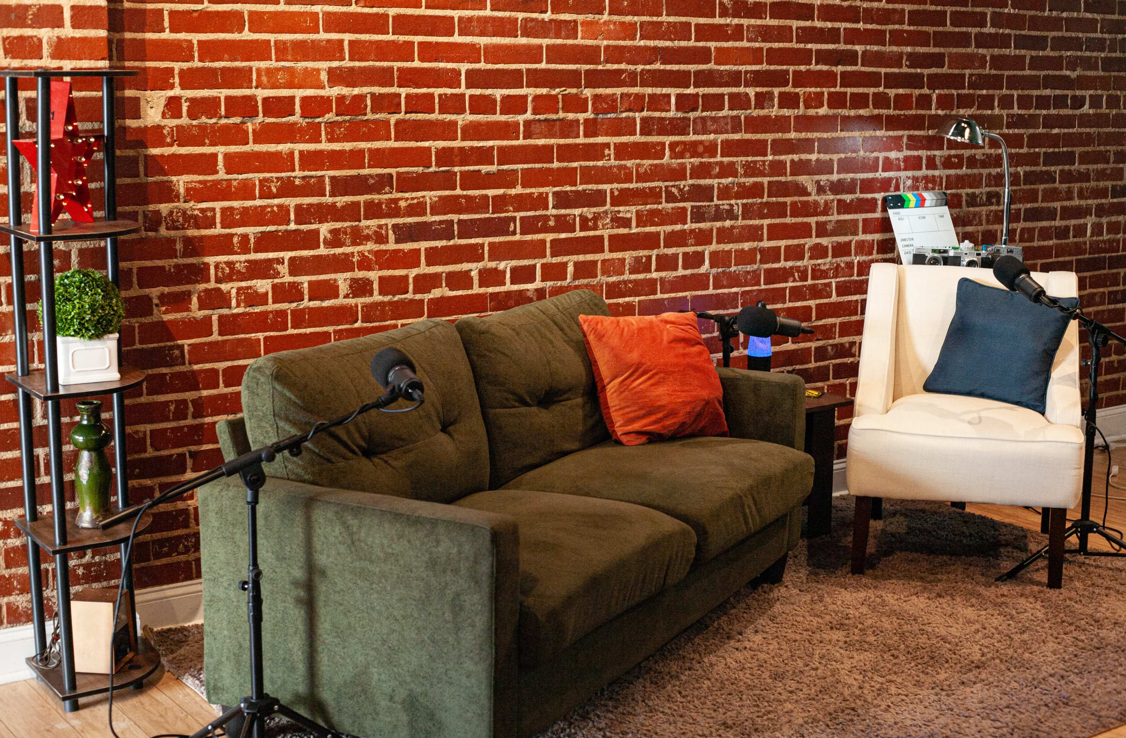 The image shows a cozy recording space featuring a green couch and a white chair next to a brick wall, with microphones positioned on stands nearby.
