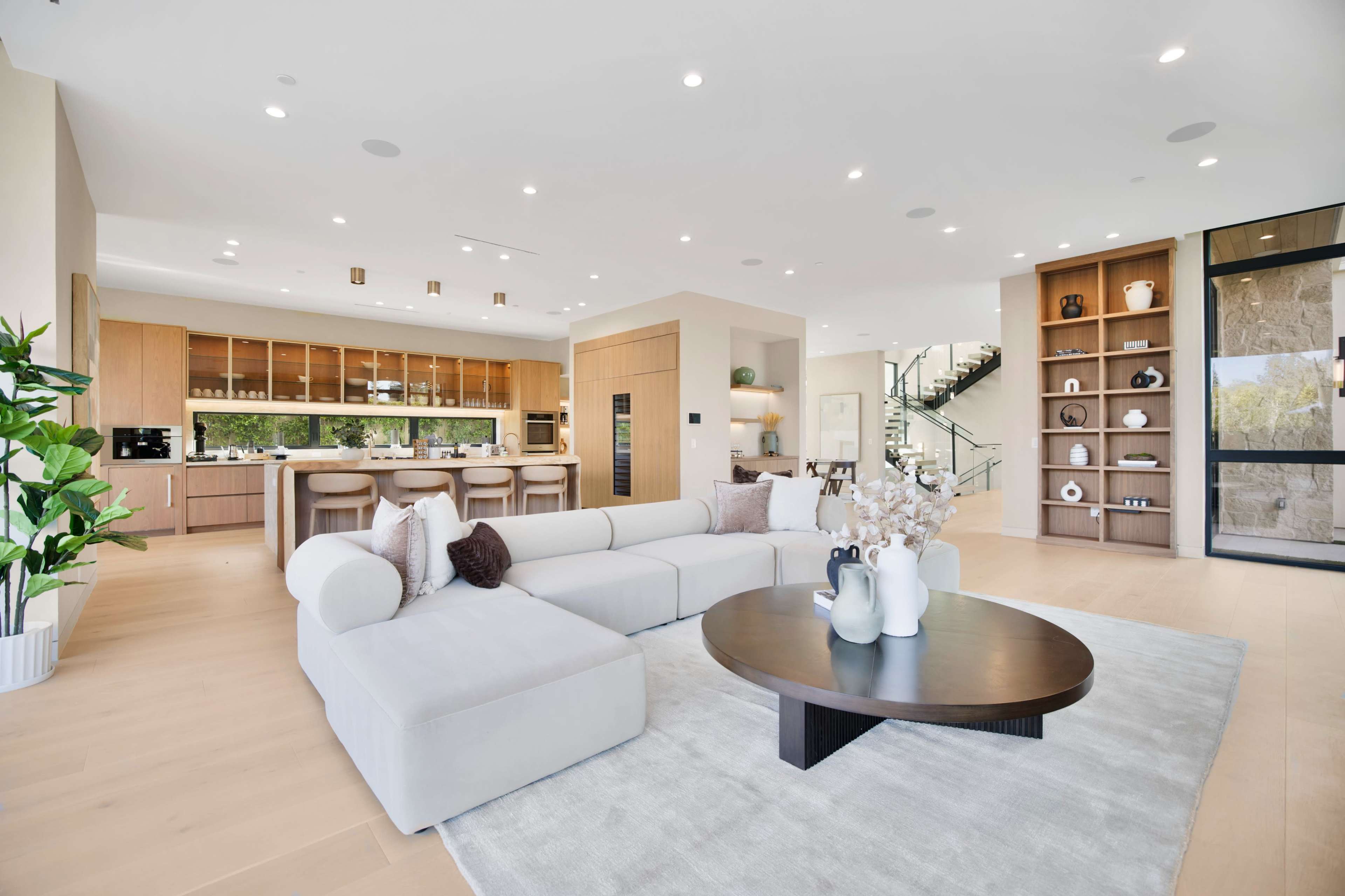 The image shows a modern open-concept living space featuring a light-colored sectional sofa, a round coffee table, a large kitchen area with wooden cabinetry, and a staircase in the background.