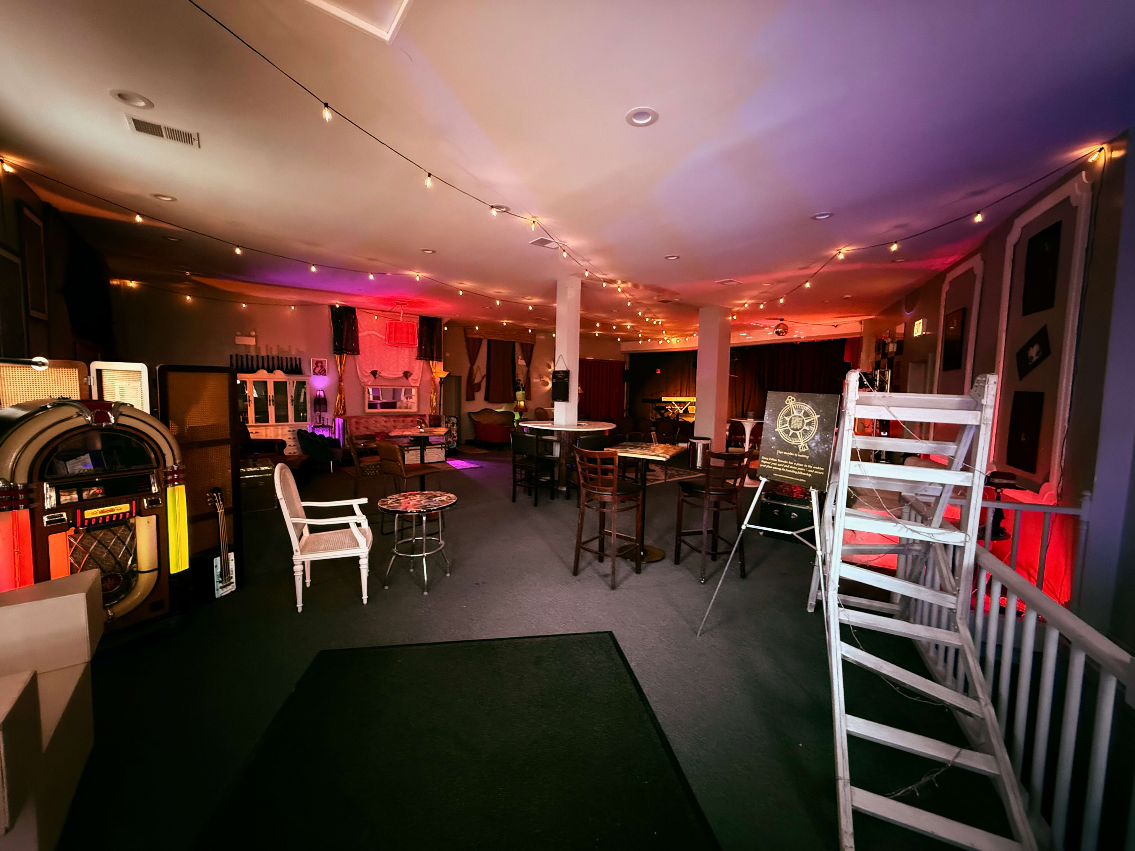 The interior of a dimly lit lounge features several tables and chairs along with a jukebox and decorative lighting.